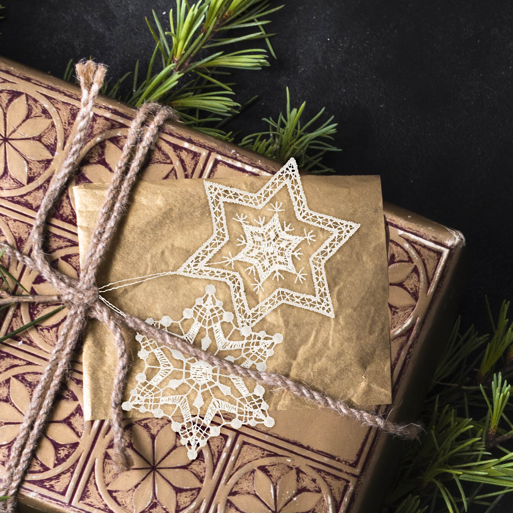 Ein goldenes Geschenk mit braunem Kordelband verpackt, darauf liegen zwei filigran gearbeitete Sterne aus Plauener Spitze im Schneestern-Design.