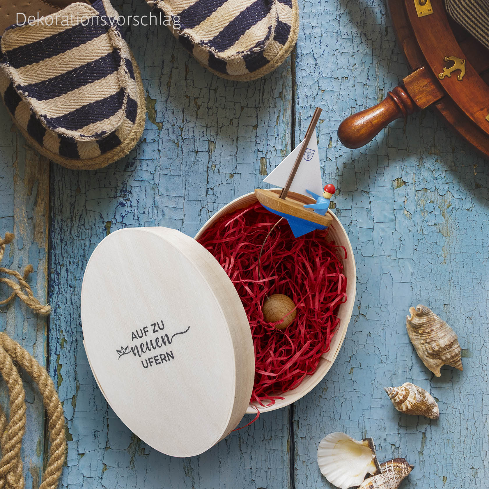 Eine runde Spandose mit rotem Füllmaterial und einem kleinen Schaukel-Segelboot darin. Der Deckel trägt die Aufschrift 'AUF ZU NEUEN UFERN'. im Dregeno Online Shop günstig kaufen