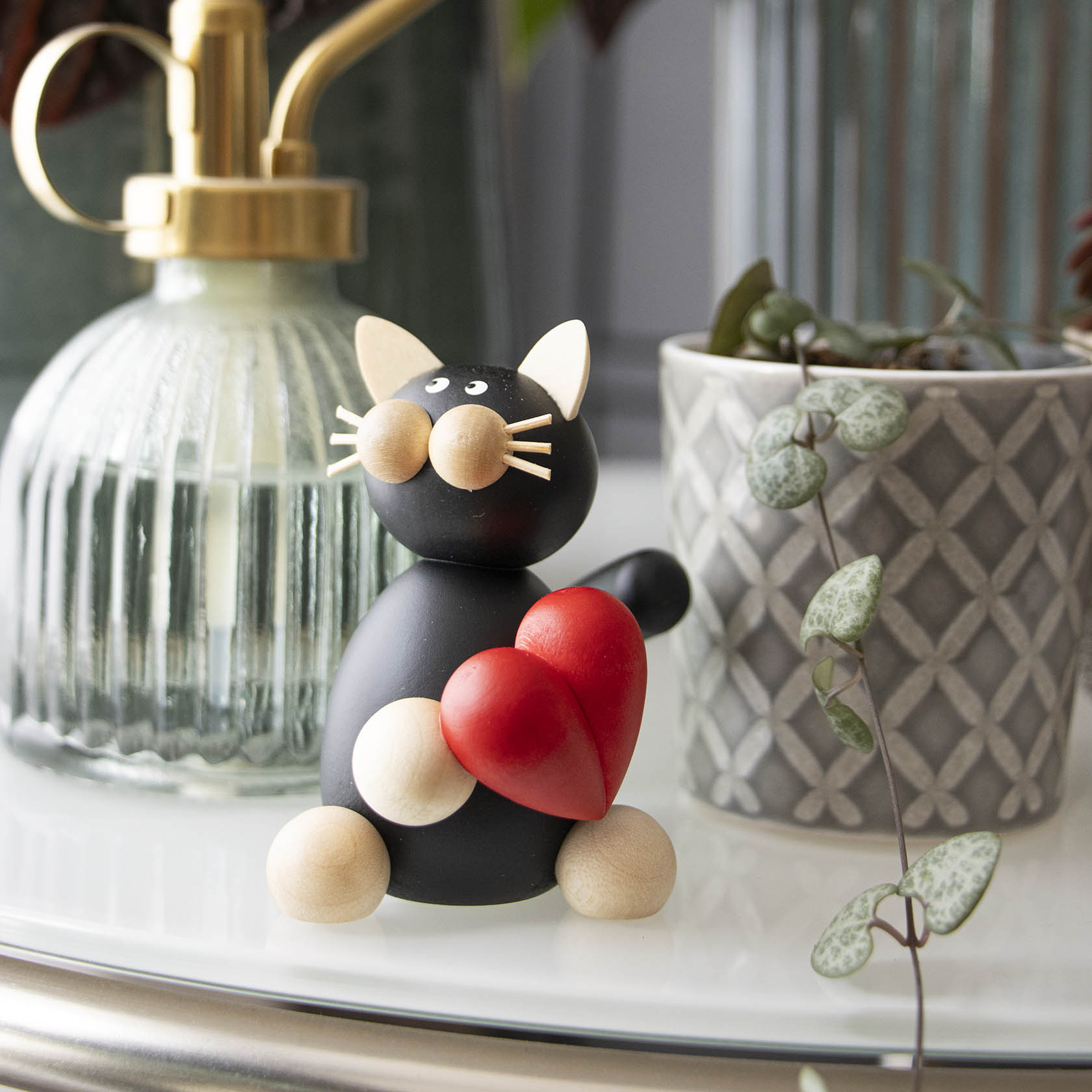 Holzfigur einer schwarzen Katze namens Hilde, die ein rotes Herz mit den Pfoten hält. Die Katze hat weiße Ohren, Schnurrhaare und eine kugelförmige Nase.