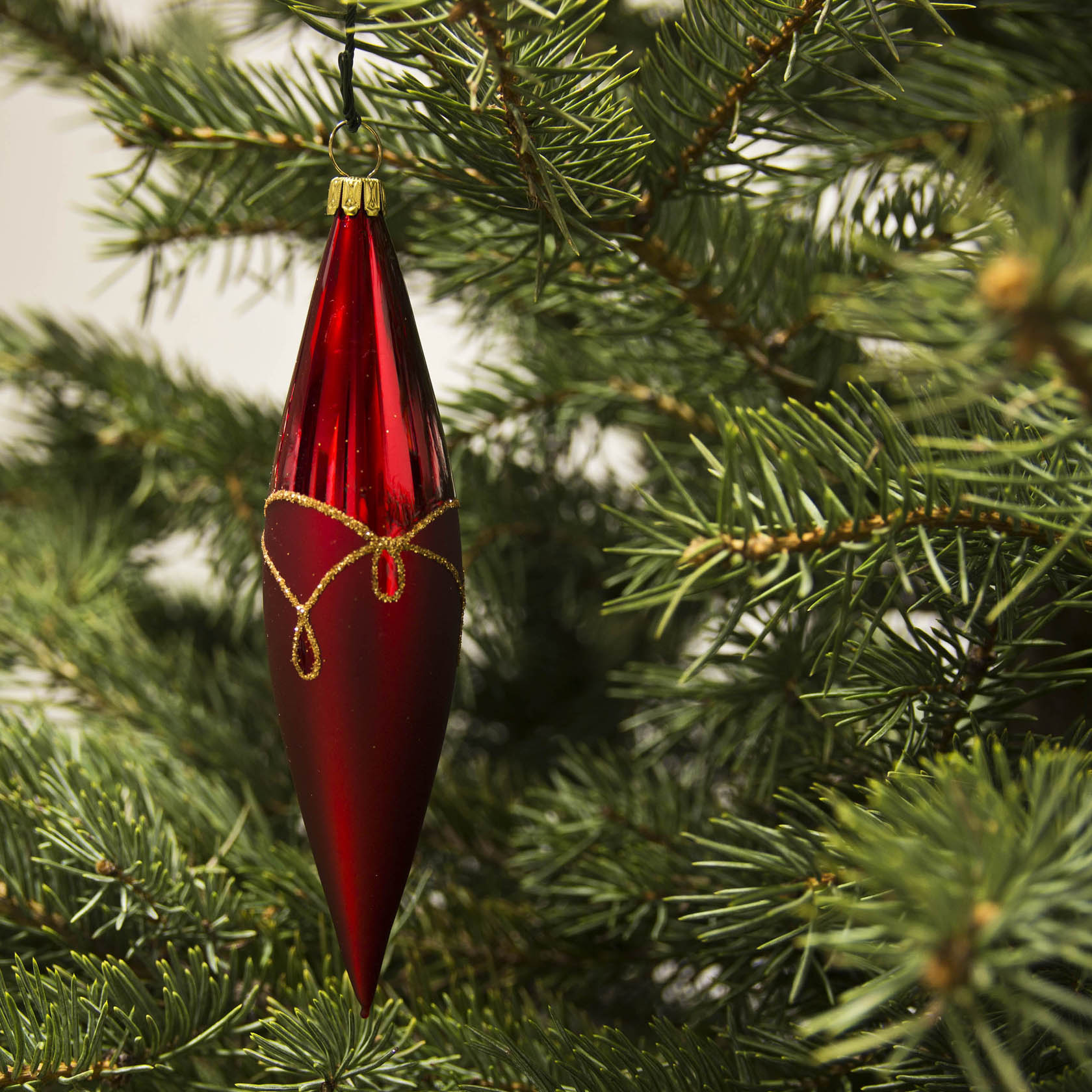 A red matte Christmas tree ornament shaped like an olive with a gold drop pattern, hanging from a branch of a Christmas tree.