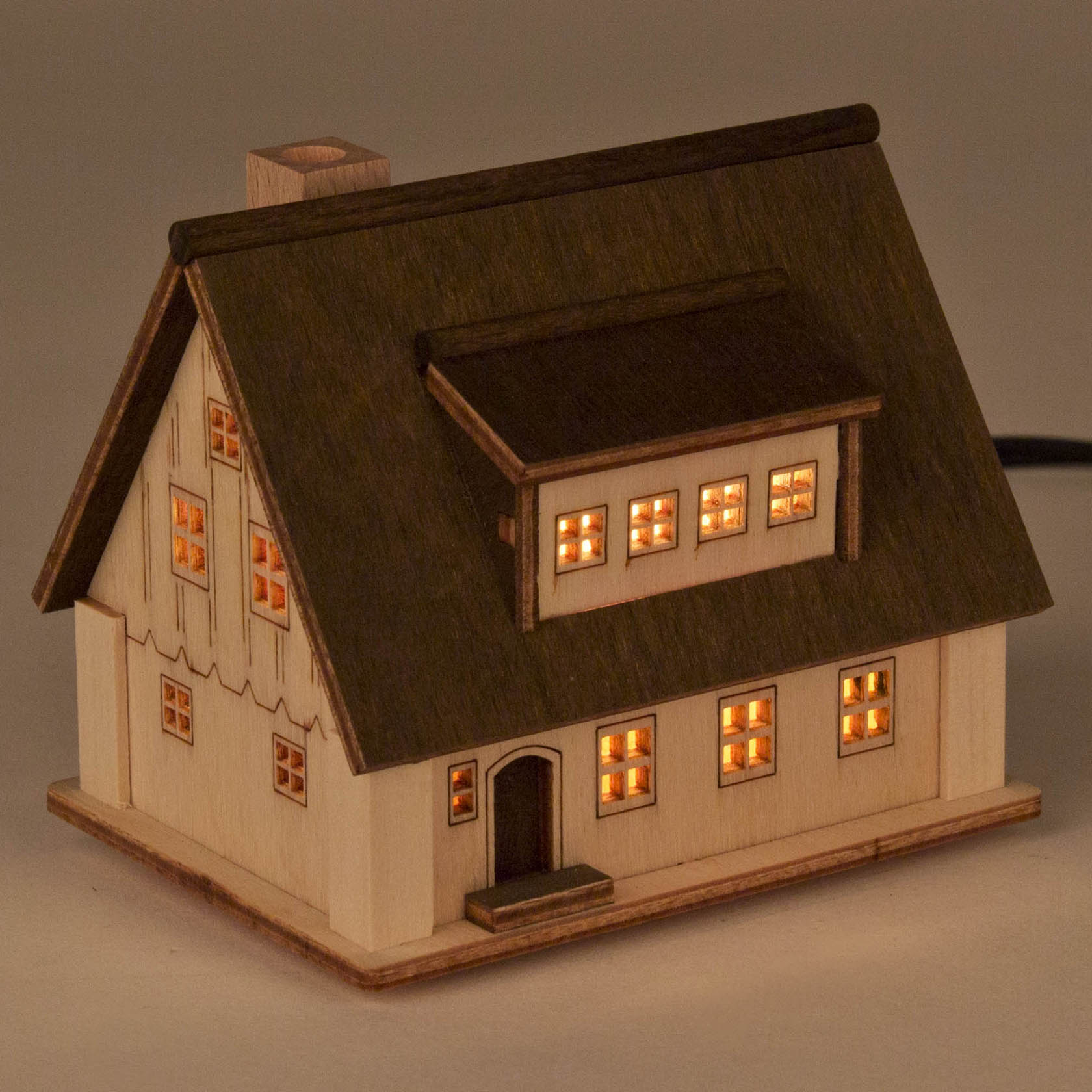 Holzmodell eines beleuchteten Hauses mit mehreren Fenstern, die warmes Licht ausstrahlen. Das Dach und die Fassade sind detailliert ausgestaltet.