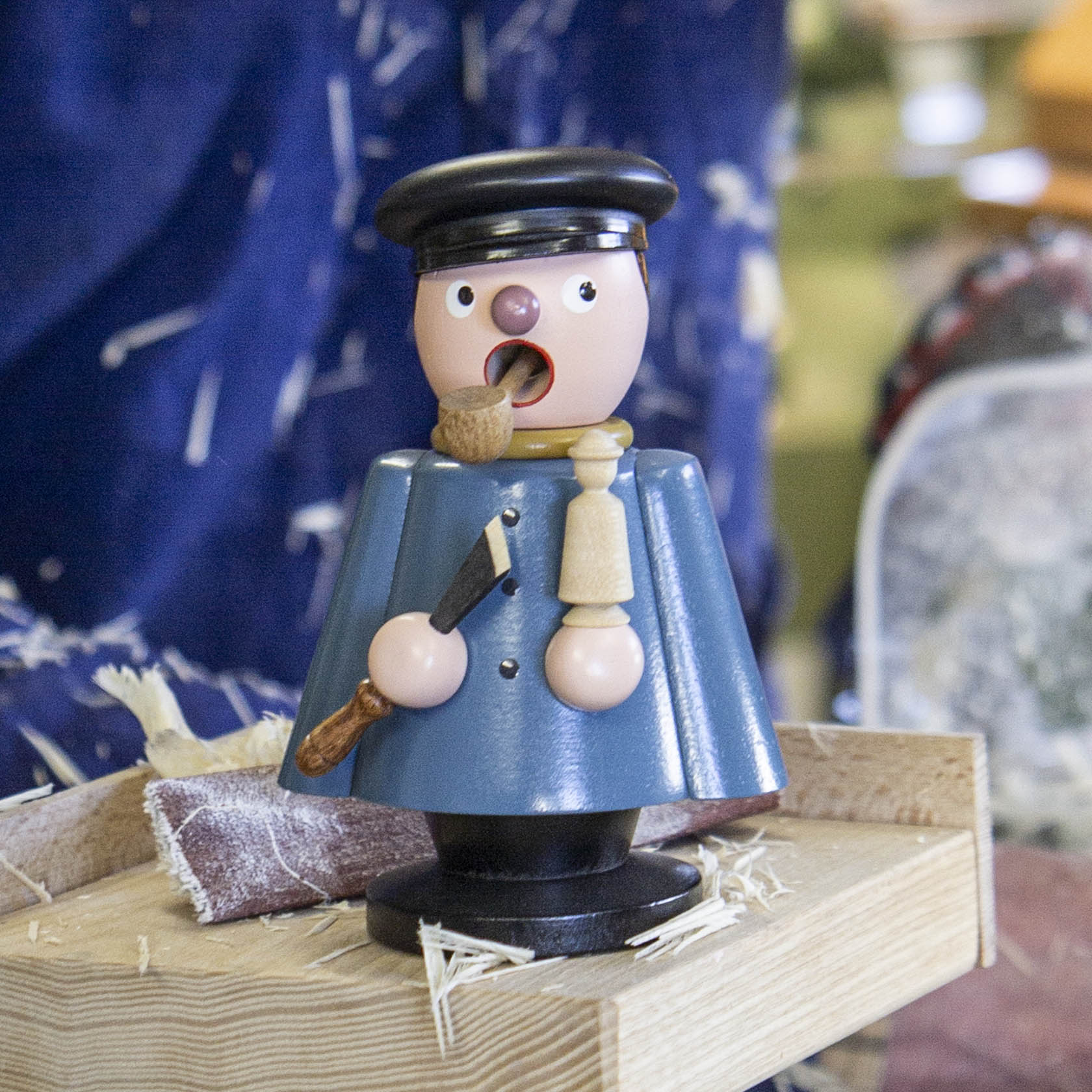 Holzfigur eines Drechslers als Räuchermann, mit Pfeife und Werkzeug in der Hand, bekleidet mit blauem Umhang und Mütze auf einem Holztisch stehend.
