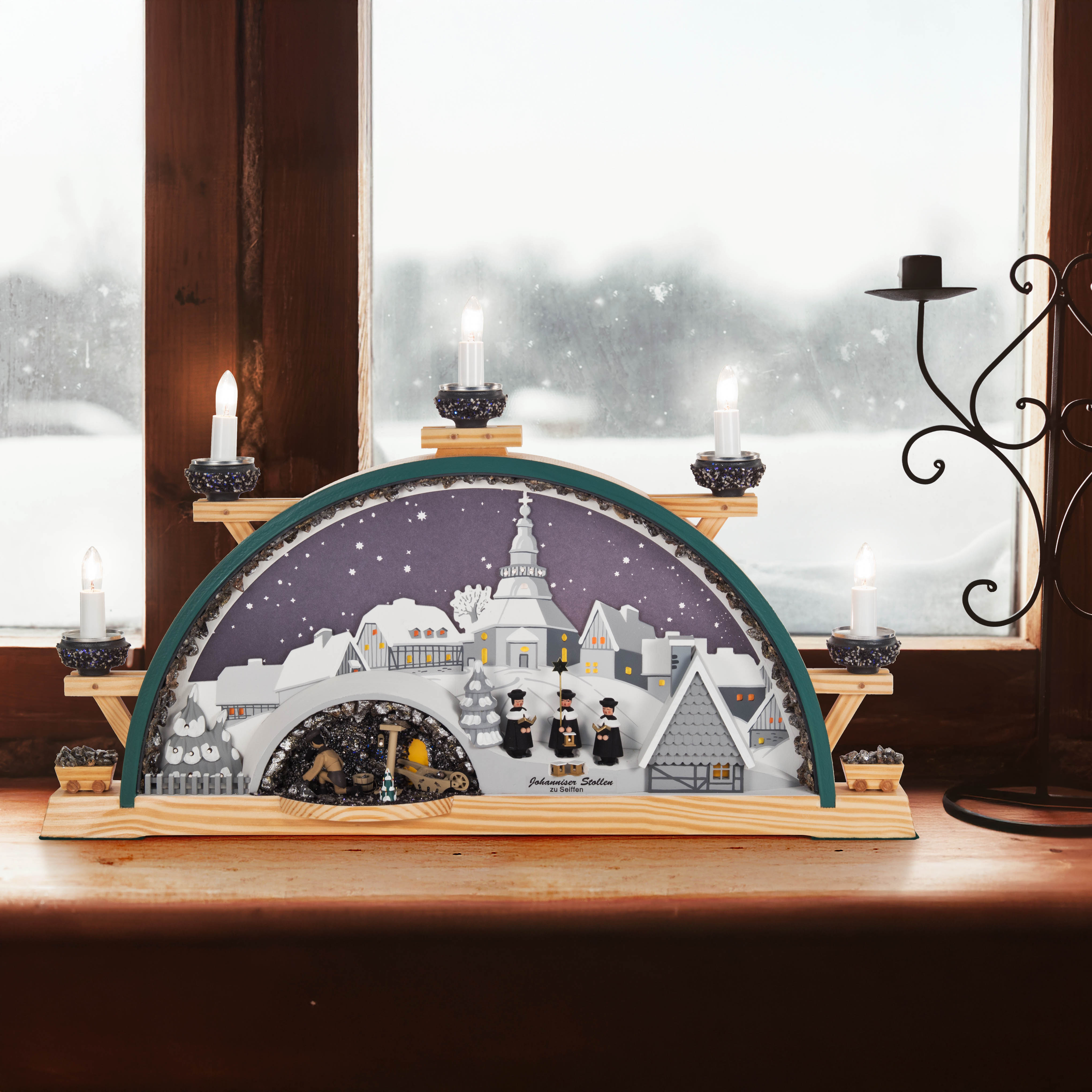 Ein elektrisch beleuchteter Schwibbogen zeigt ein winterliches Seiffener Dorf mit Kirche, Kurrende-Sängern und Bergwerk. Sechs Kerzenleuchten veredeln die Szenerie.