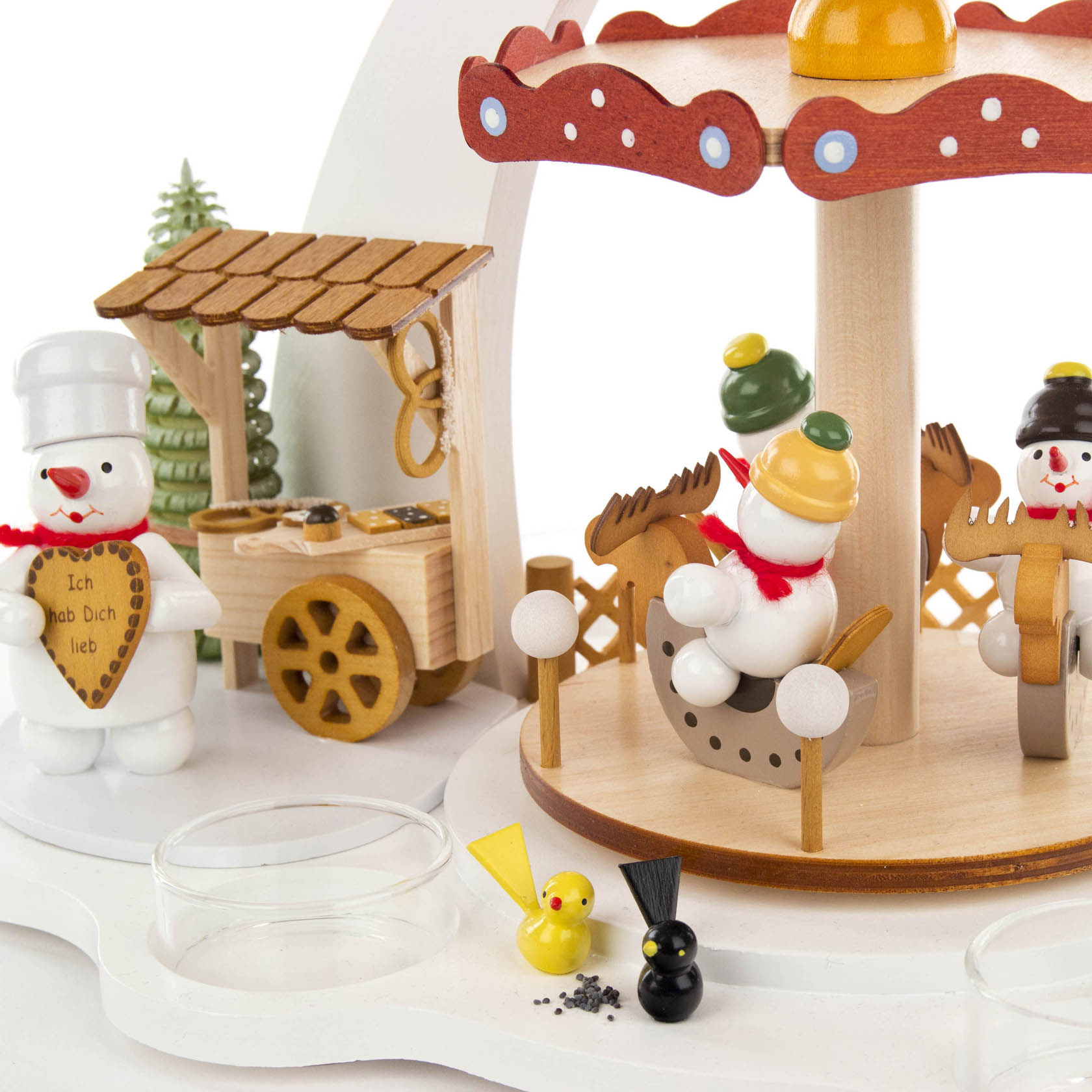 Holzschneemann im Kochanzug hält ein Lebkuchenherz neben einer Miniaturdrehkarussell mit weiteren Schneemännern, die auf Schaukelpferden sitzen.