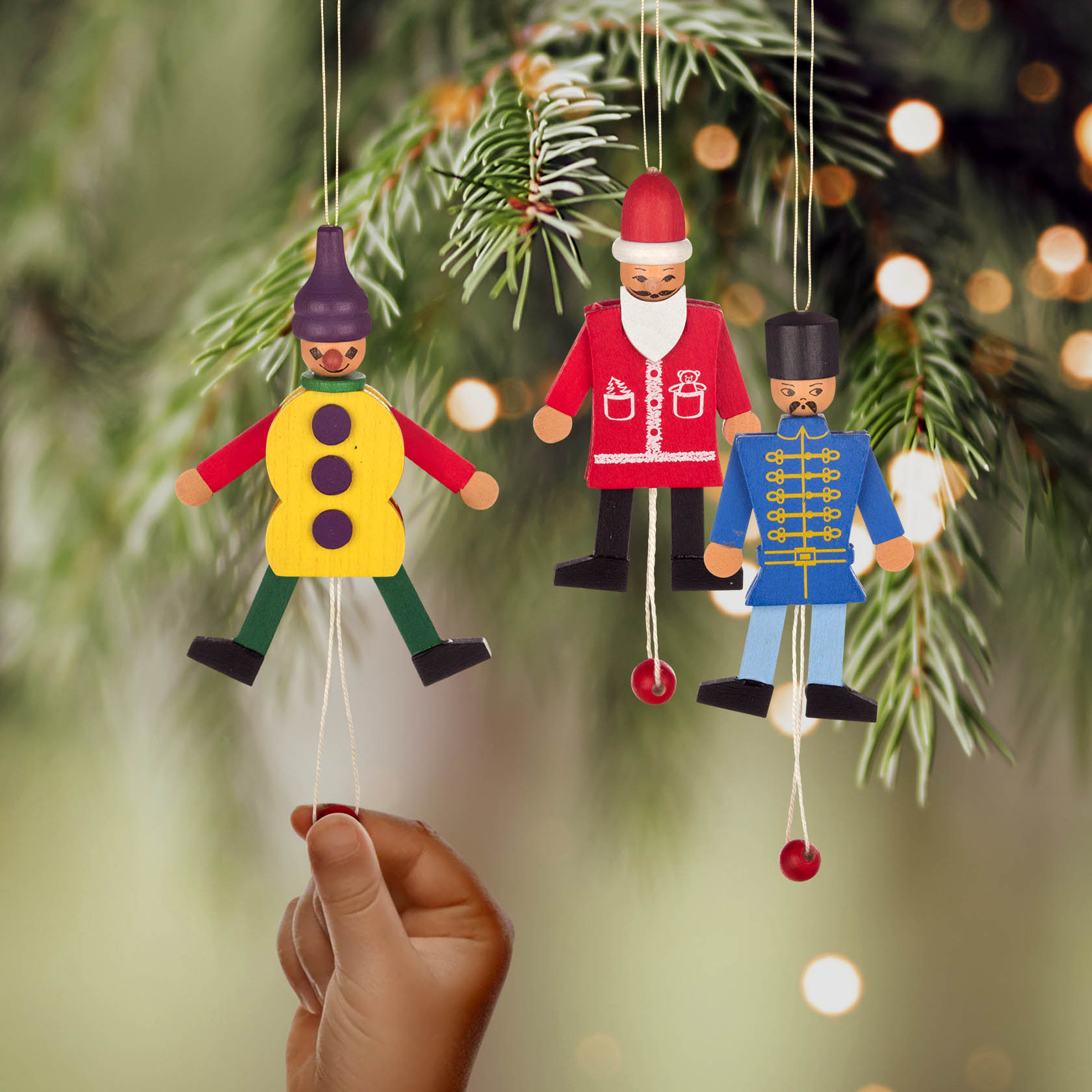Hand zieht an einem Holz-Hampelmann in Soldatenform, der an einem Weihnachtsbaum hängt. Zwei weitere Hampelmänner, ein Weihnachtsmann und ein Harlekin, daneben.