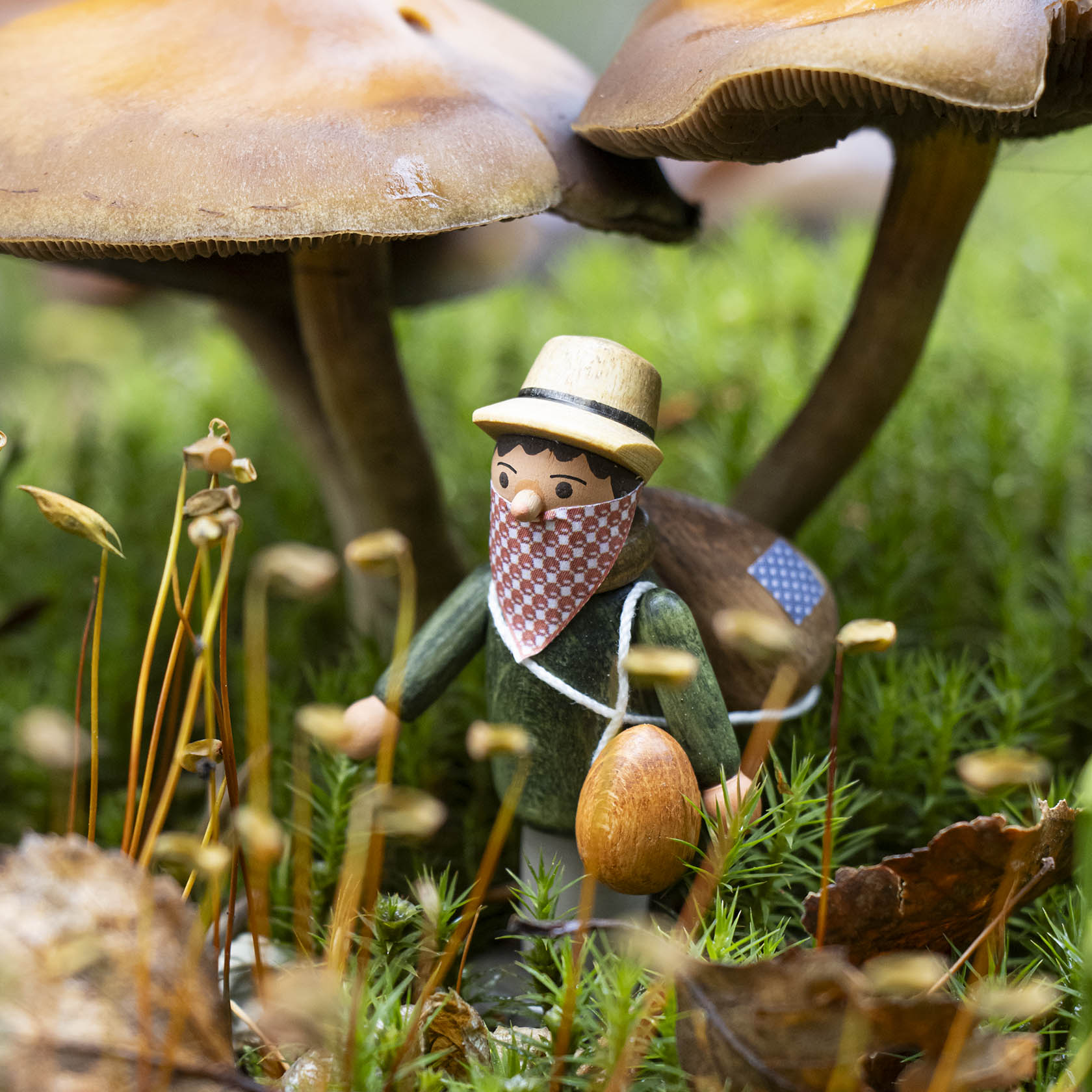 Kleine Holzfigur eines Mannes mit Hut und rotem Halstuch, der einen Pilz in der Hand hält. Er steht zwischen hohen Pilzen auf einer Wiese.