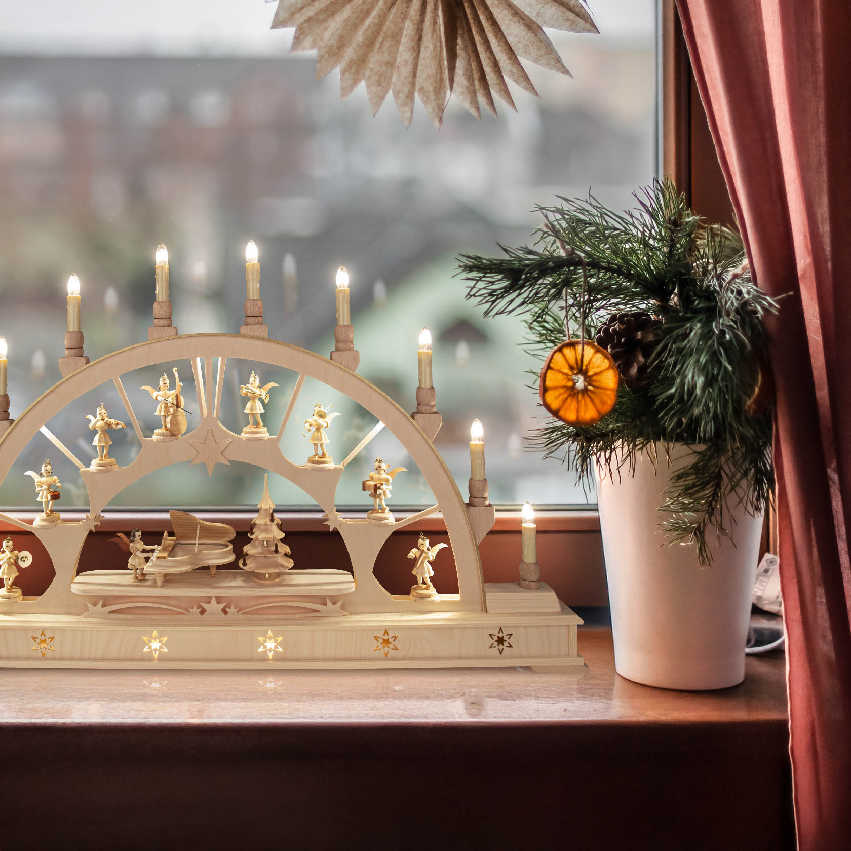 Ein beleuchteter Schwibbogen mit Sammelfiguren und LED-Lichtern steht auf einer Fensterbank neben einem dekorierten kleinen Topf mit Tannenzweigen und getrockneter Orangenscheibe.