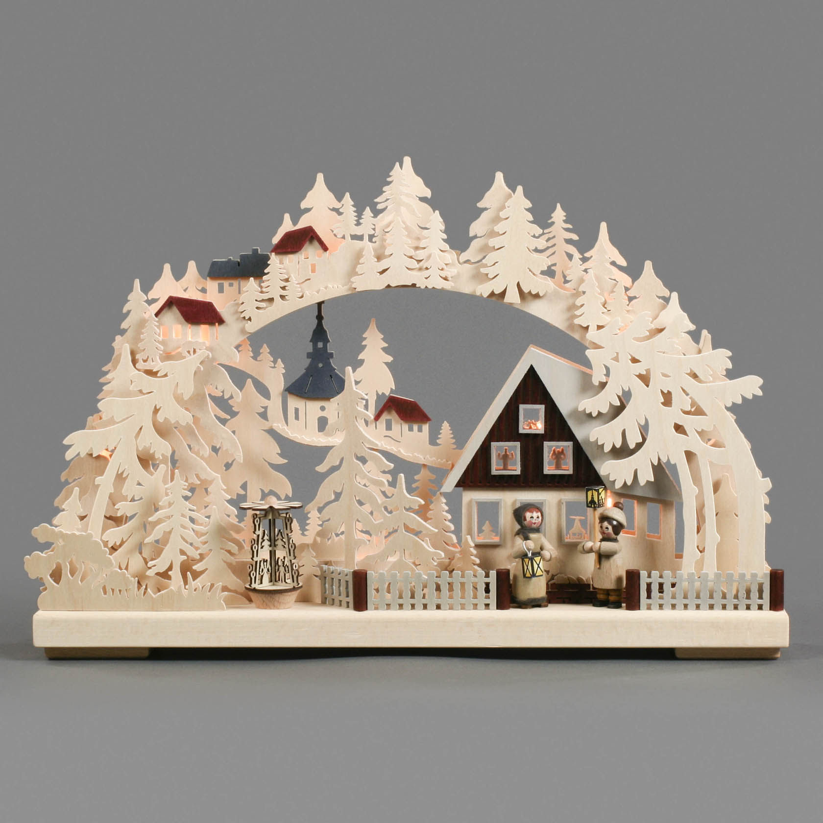 Holz-Schwibbogen mit geschnitzten Tannenbäumen und Figuren, die ein Haus mit Zaun sowie eine Kirche im Hintergrund zeigen. Die Szene ist elektrisch beleuchtet.