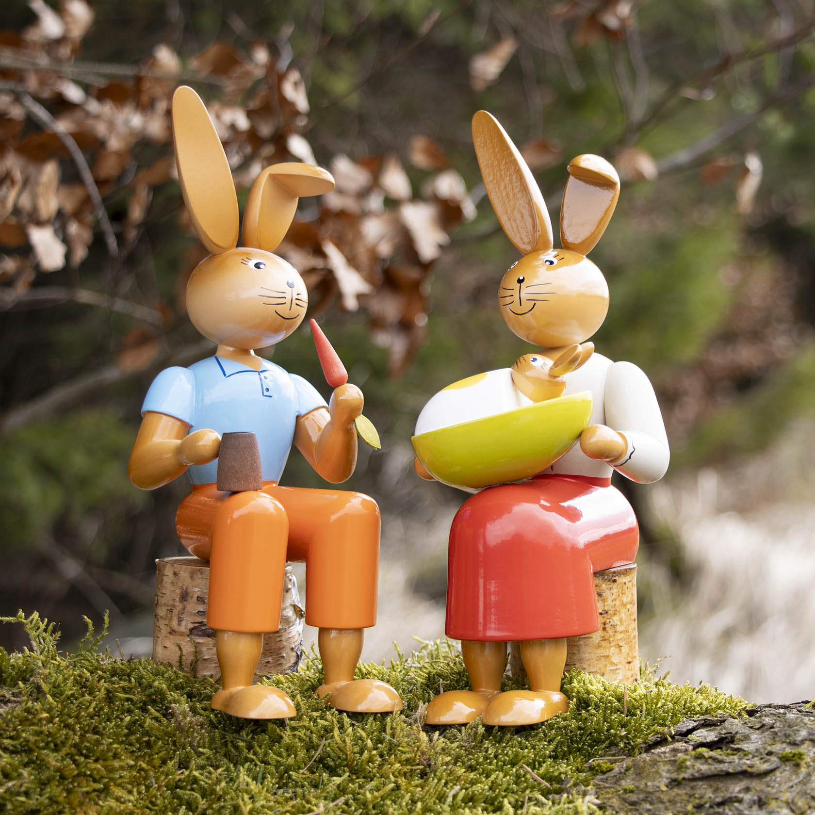 Zwei Hasenfiguren aus Holz, sitzend auf kleinen Baumstämmen. Links ein Hase mit blauer Figur und Karotte, rechts Häsin mit rotem Rock und grünem Gefäß.