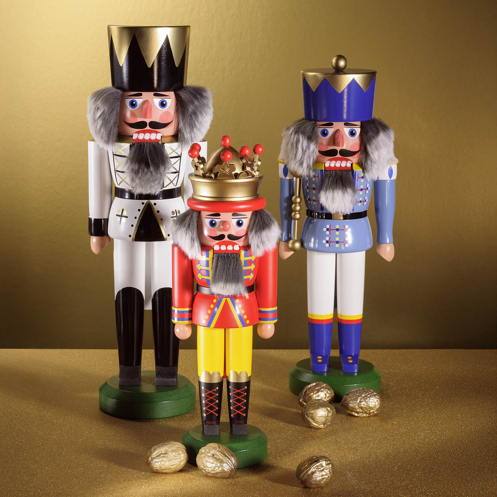 Three wooden nutcracker figures: a tall king in white and black with a gold crown, a king in red and yellow with a gold crown, and a king in blue and white with a blue crown, standing on a sparkly golden surface surrounded by nuts.