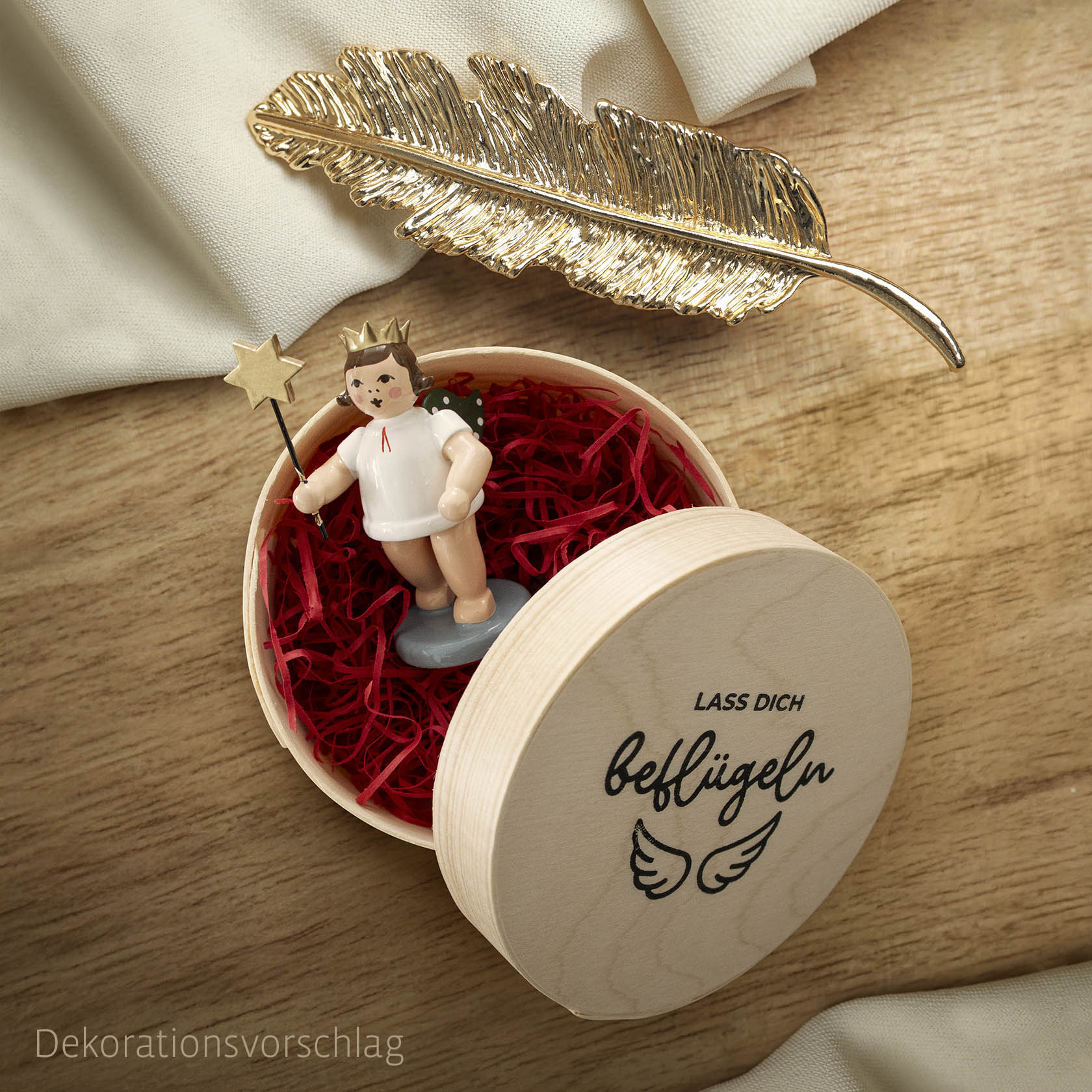 Ein Engel mit Krone und roter Schleife hält einen Sternstab, platziert in einer runden Holzspandose mit Aufschrift 'Lass dich beflügeln'. im Dregeno Online Shop günstig kaufen