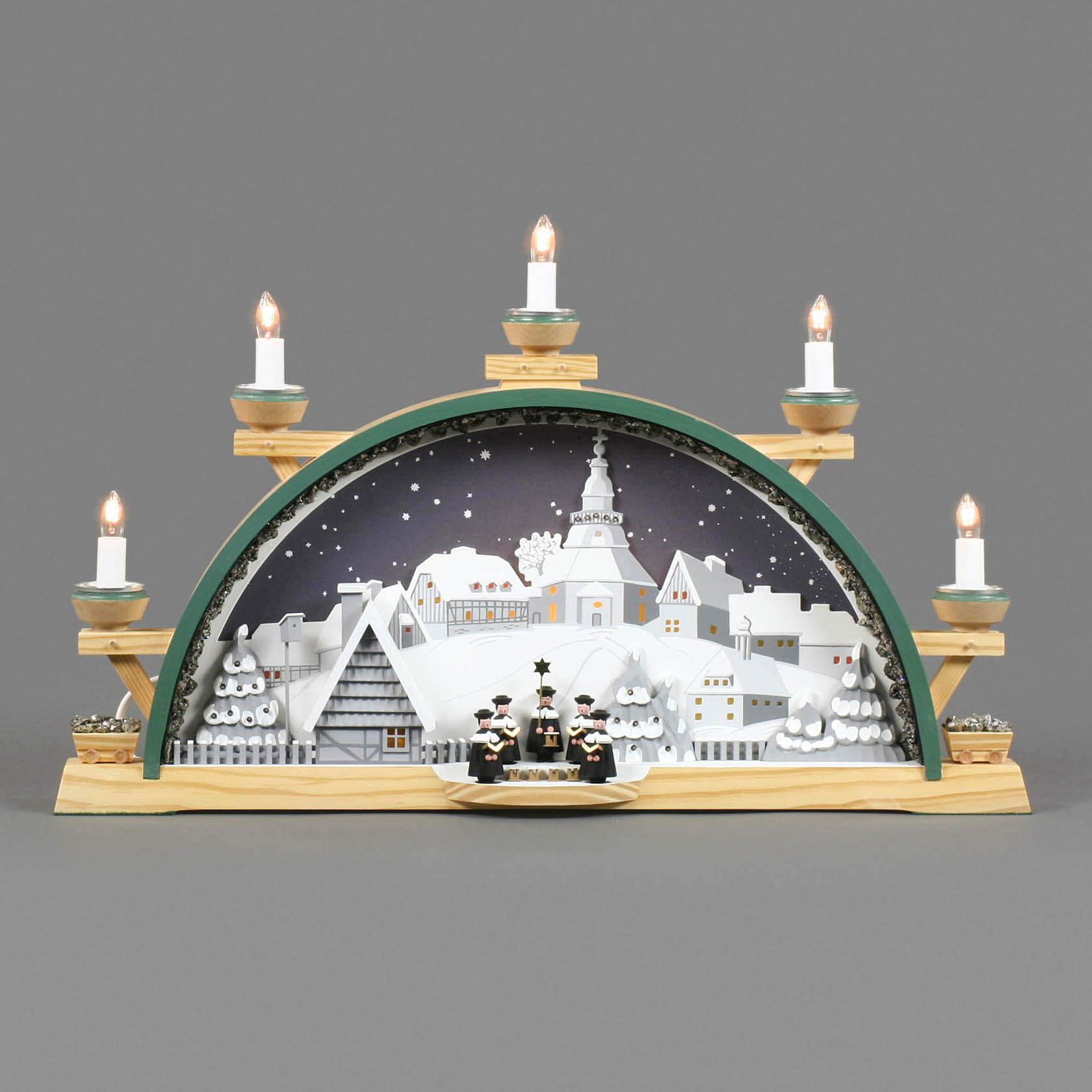 Elektrisch beleuchteter Schwibbogen mit Seiffener Dorf und Kurrendefiguren. Im Hintergrund verschneite Gebäude und eine Kirche, im Vordergrund drei Kurrendesänger.