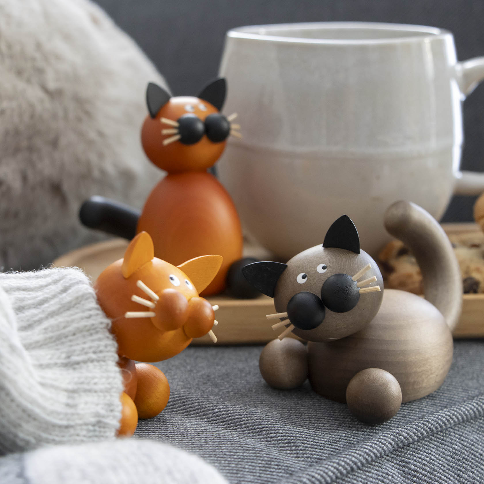 Drei dekorative Holzfiguren von Katzen, eine grau-schwarz und zwei in Orange, sitzen auf einem grauen Stoff neben einem Becher.