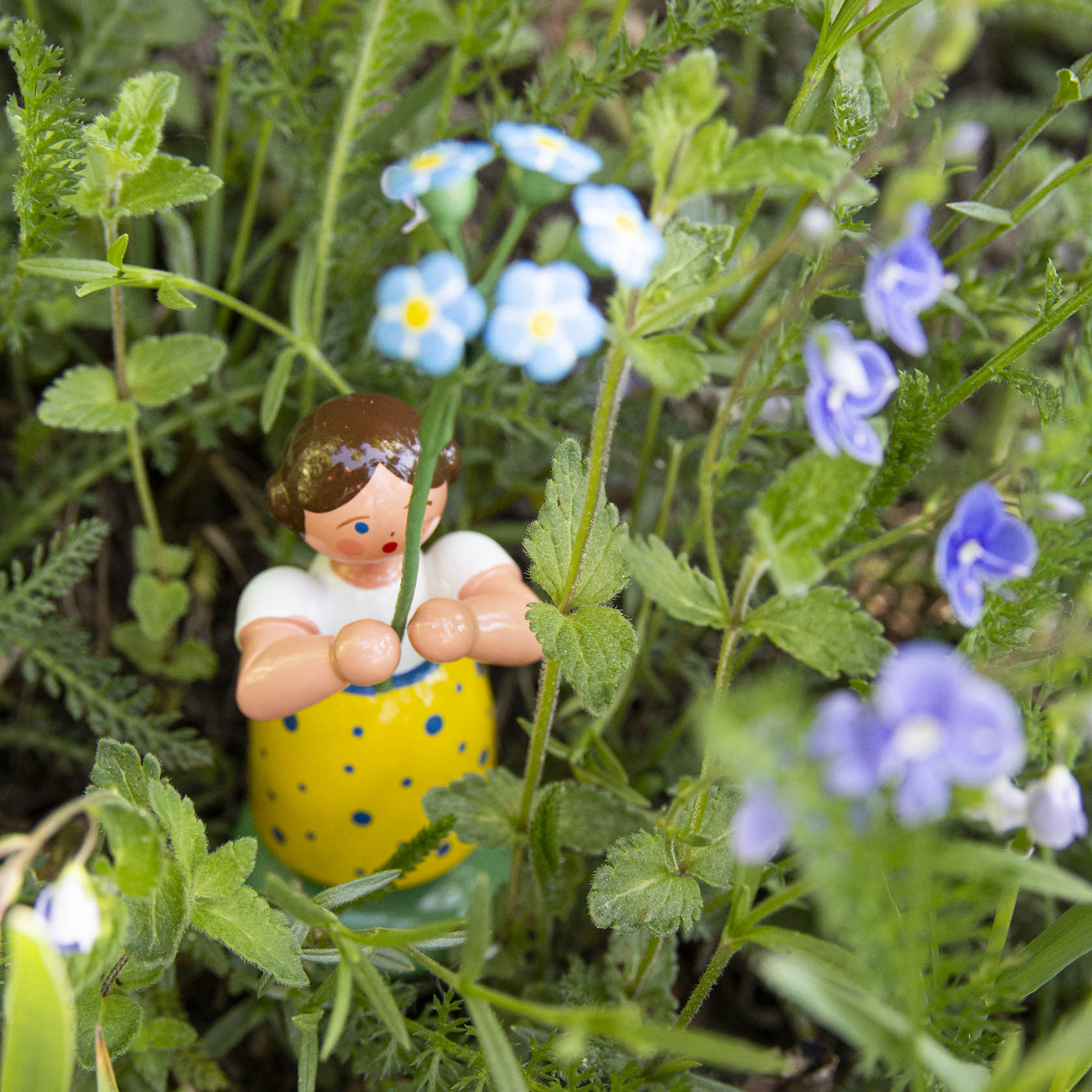 Holzfigur eines Mädchens in gelbem Kleid, das einen Strauß Vergissmeinnicht hält, umgeben von grünen Blättern und blauen Blumen.