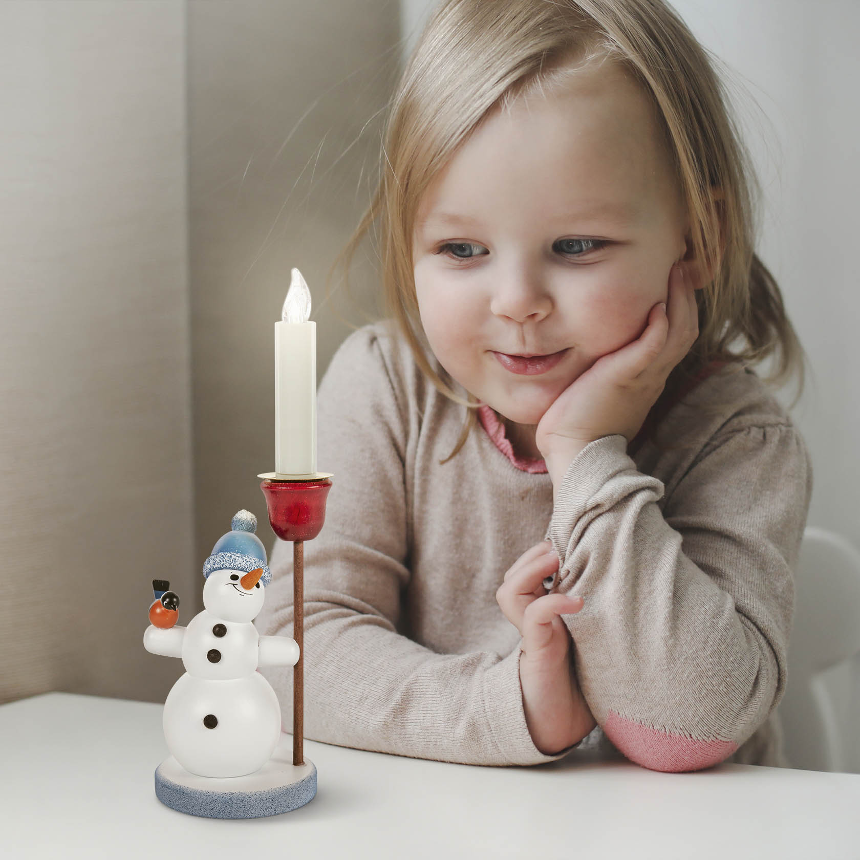Ein kindliches Mädchen lächelt einen dekorativen Schneemann an, der einen blauen Pudelmütze trägt und einen Kerzenhalter mit brennender Kerze hält.