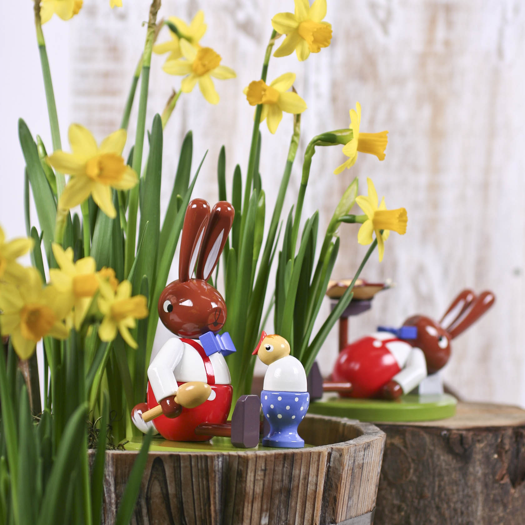 Zwei große hölzerne Hasenfiguren in traditioneller Kleidung, eine sitzend mit einem bemalten Ei in einem blauen Eierbecher, umgeben von gelben Narzissen.