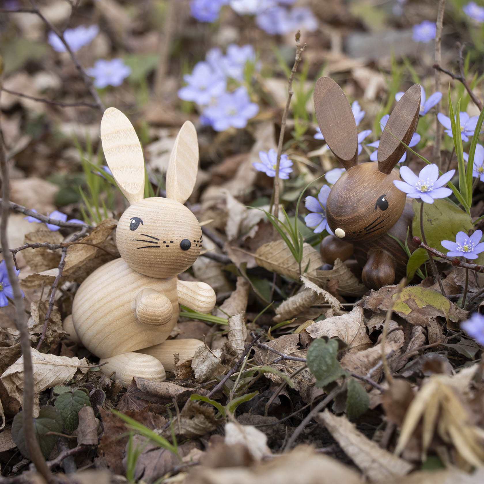 Zwei Holzosterhasen, ein heller und ein dunkler, sitzen zwischen violetten Blumen auf vertrocknetem Laub.