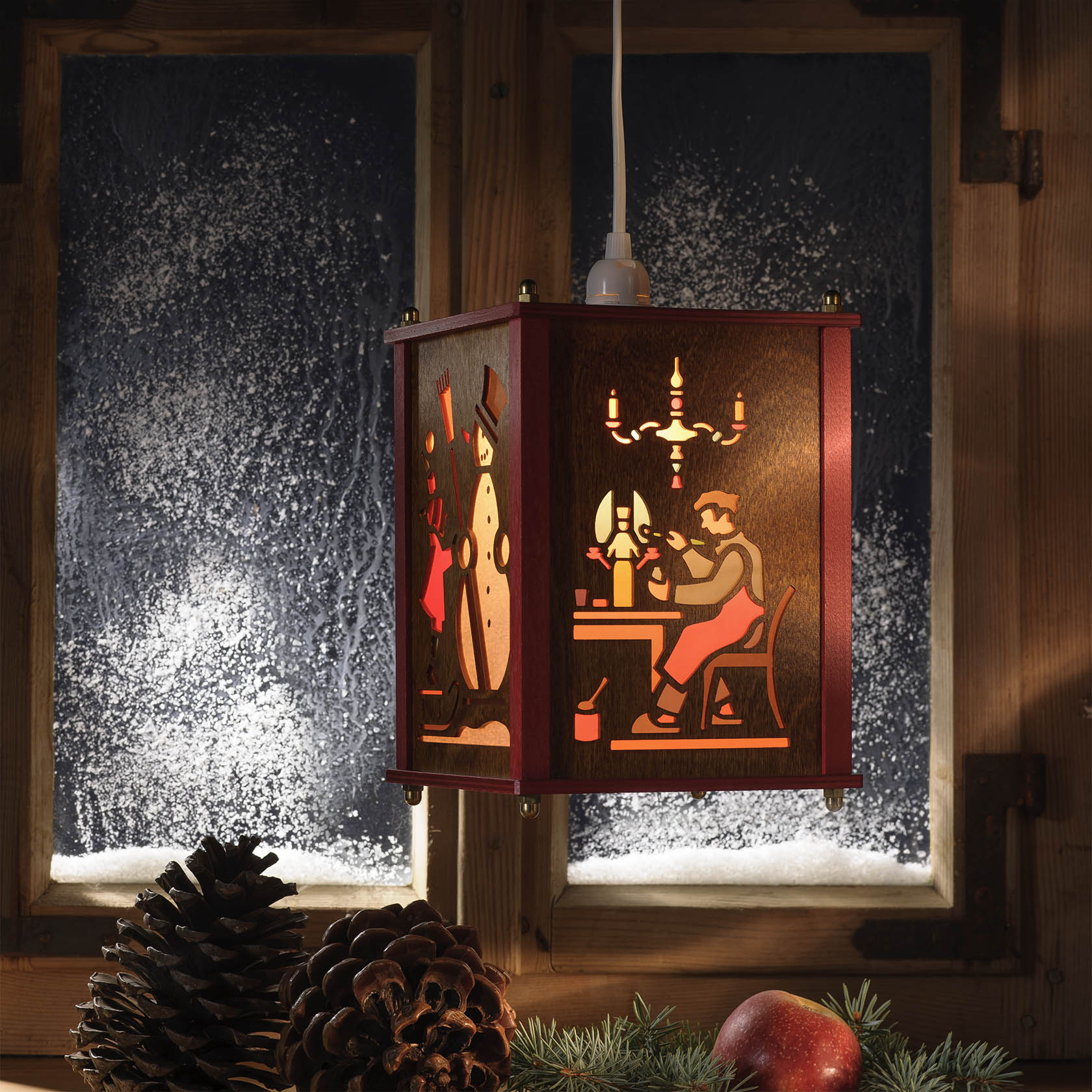 Beleuchtete Laterne mit Scherenschnitt von Weihnachtsmann, Schneemann und Spielzeugmacher. Im Hintergrund ein mit Schnee bedecktes Fenster.