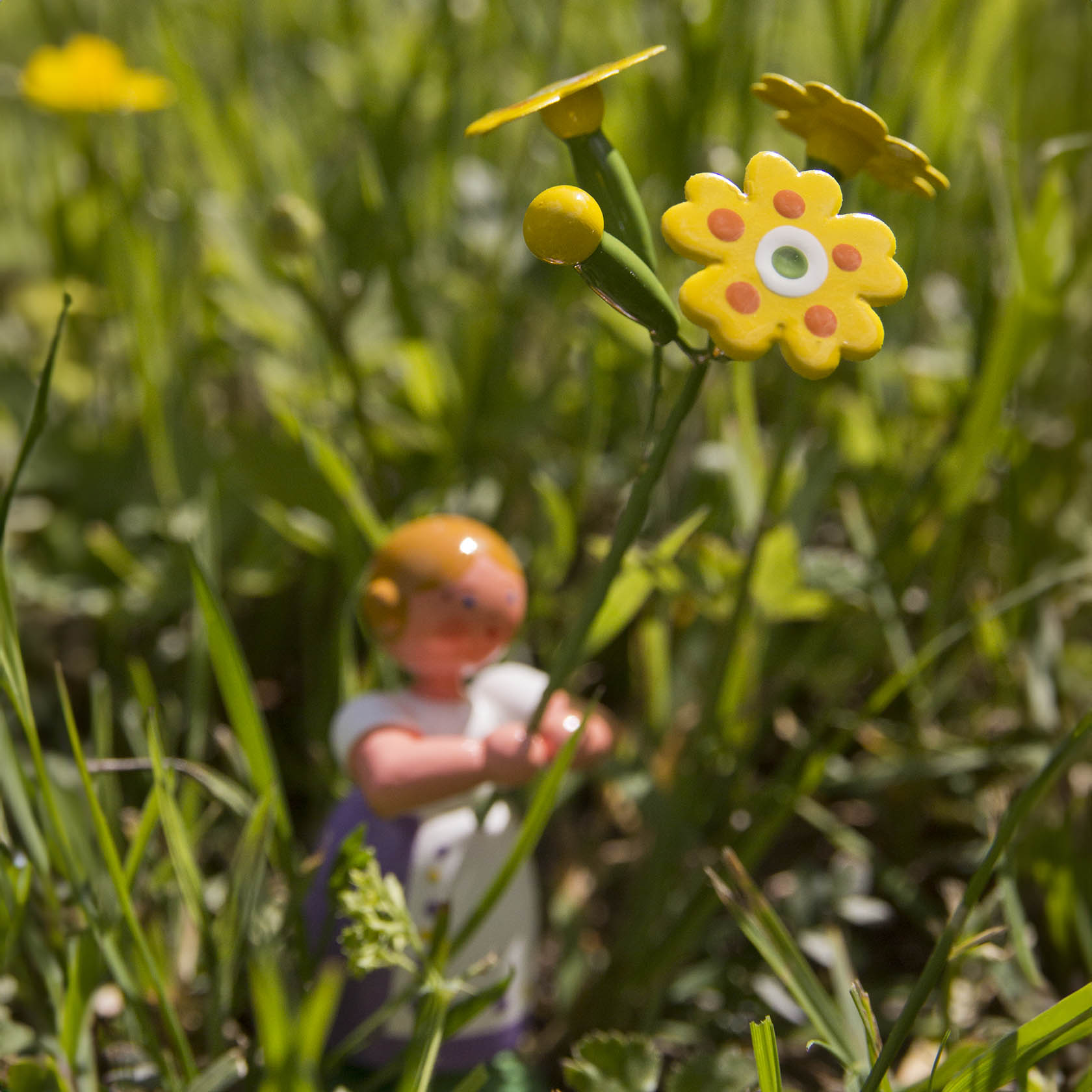 Eine Spielzeugfigur eines Mädchens in einem weißen Kleid hält eine kunststoffgelbe Blume mit Himmelschlüssel in einer grünen Wiese.