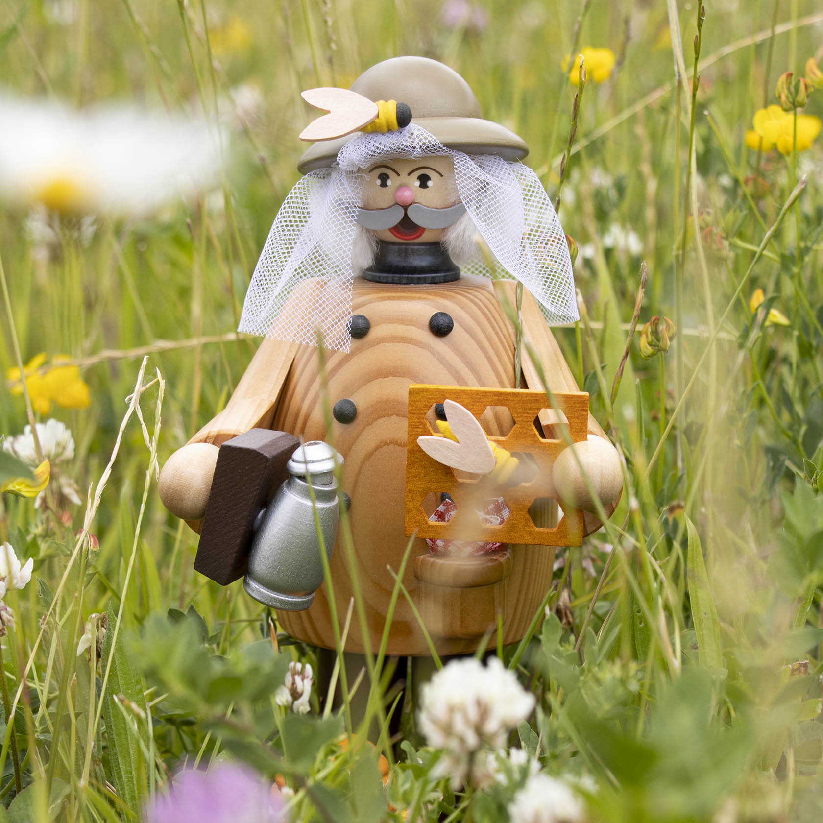 Ein hölzerner Räuchermann in Form eines Imkers steht in Wiese, trägt Hut und Schleier, hält Bienenwabe und Smoker.