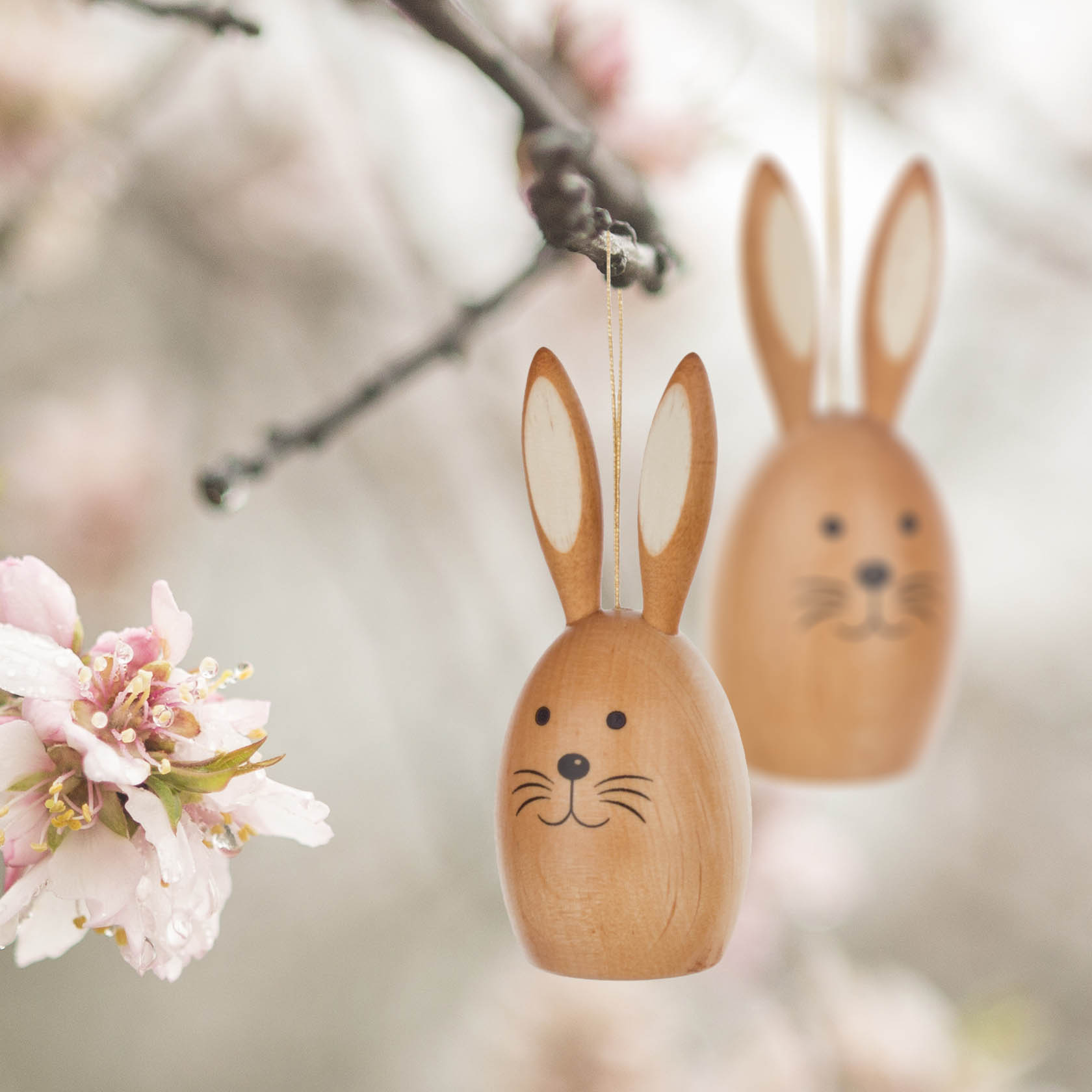 Zwei hölzerne Osterhasen-Dekorationen mit bemalten Gesichtszügen und aufgemalten Ohren hängen an einem Ast neben blühenden Blumen.