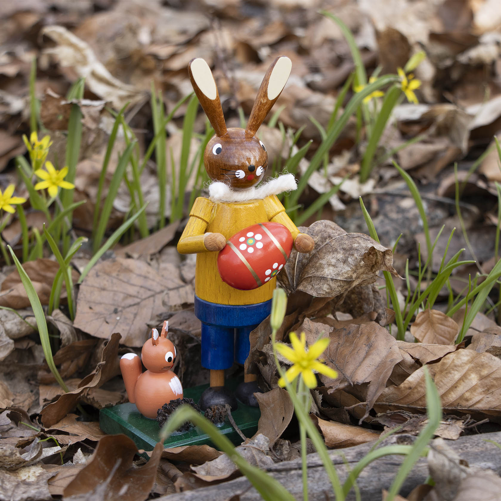 Eine hölzerne Figur eines Osterhasen in gelbem Pullover, hält ein rotes Ei mit weißen Punkten. Eine kleine Eichhörnchenfigur sitzt daneben.