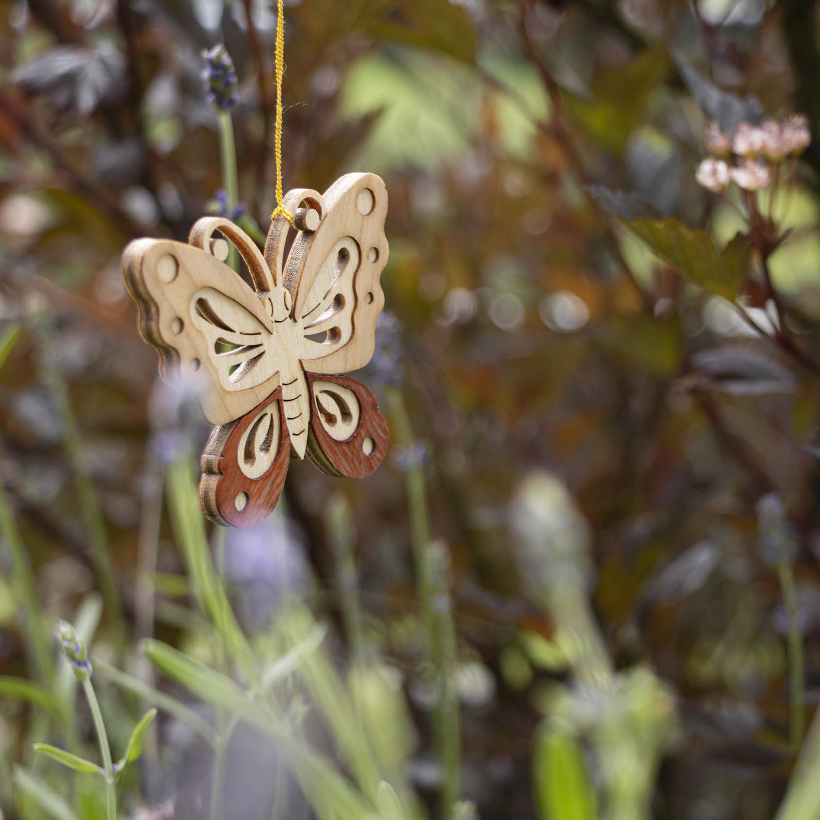 Holzschmetterling mit filigranen Details hängt an einem gelben Faden inmitten grüner Vegetation.