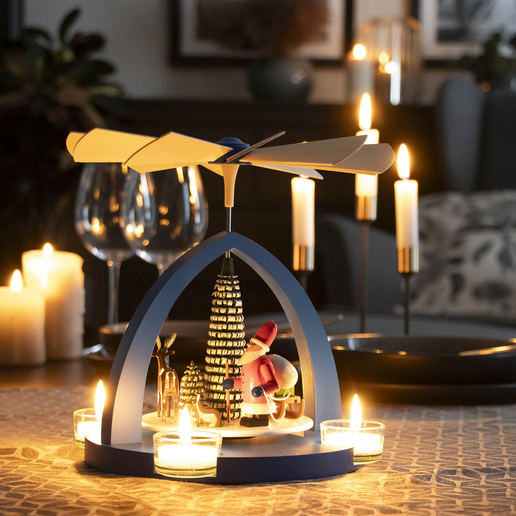 Blaue Weihnachtspyramide für Teelichter mit Weihnachtsmann-Figur und Waldtieren unter einem drehbaren Flügelrad, beleuchtet durch brennende Kerzen.