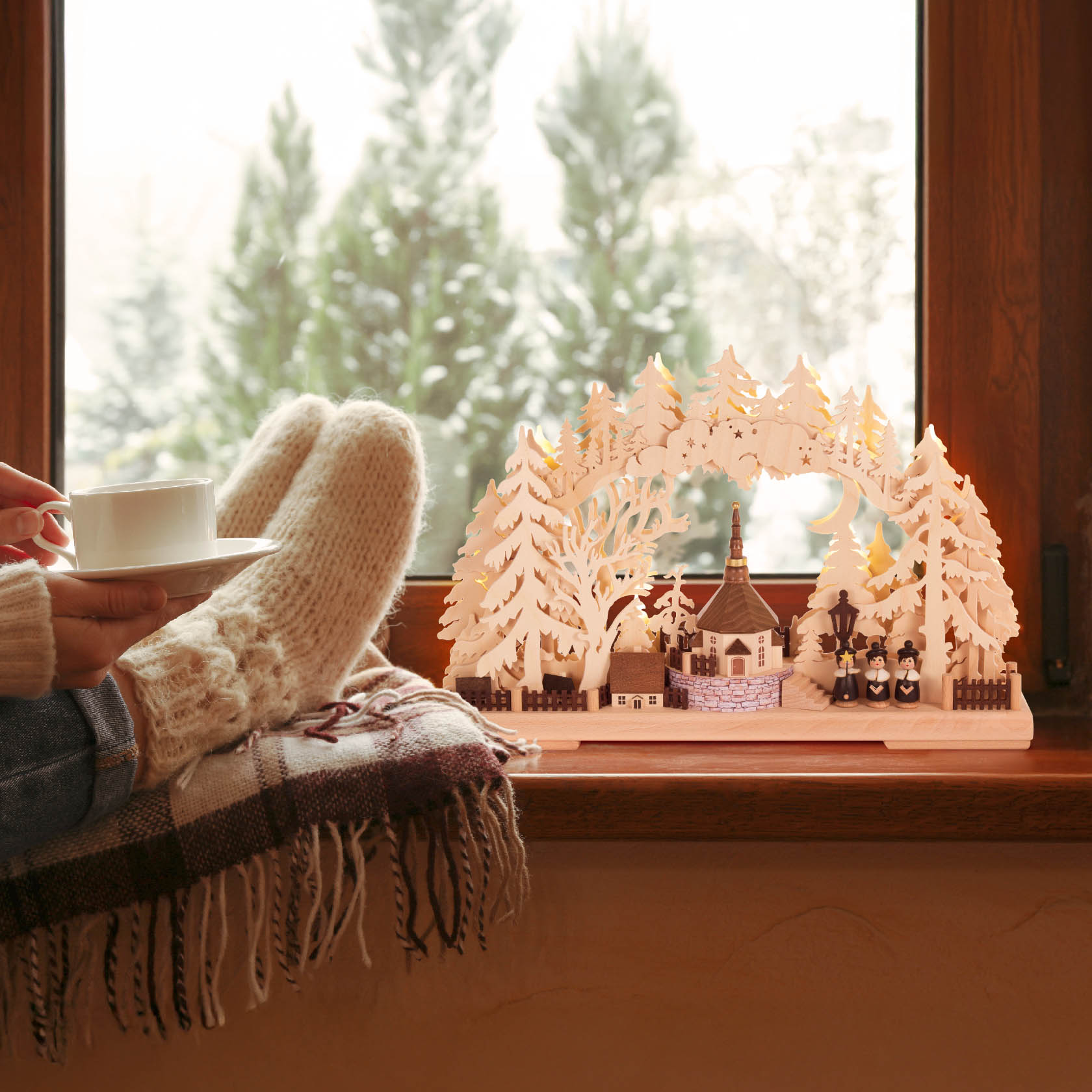 A small electric illuminated Schwibbogen showing a village church scene with surrounding trees. A person with cozy socks holds a teacup near a window.