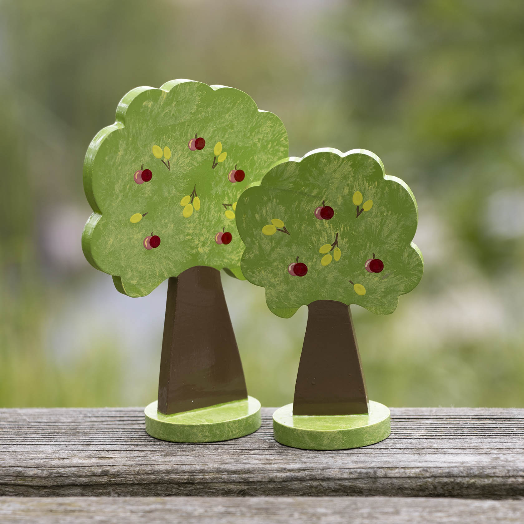 Zwei kleine Spielzeug-Apfelbäume aus Holz. Der linke Baum ist etwas größer, beide haben bemalte rote Äpfel und gelbe Blätter auf ihrer Krone.