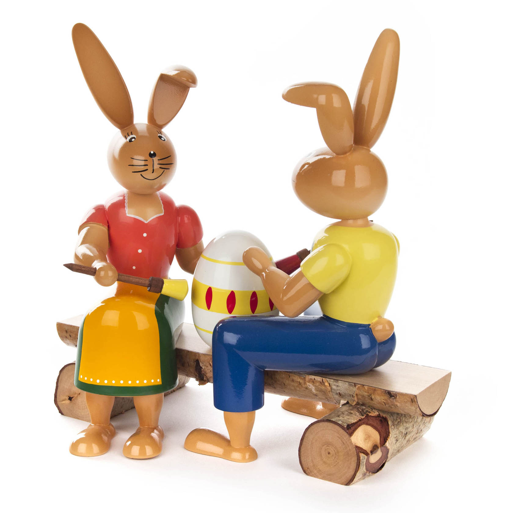 Zwei Holzhasenfiguren sitzen auf einer Holzbank. Beide halten Pinsel und bemalen gemeinsam ein großes Osterei.
