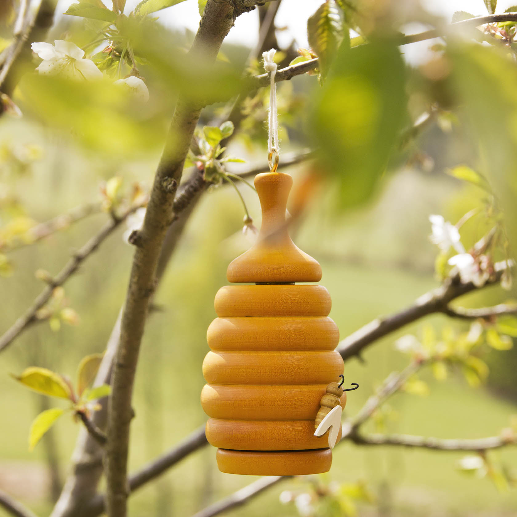 Holzbienenstock mit Räucherfunktion, der an einem Ast hängt. Er ist aus hellem und dunklem Holz gefertigt und hat eine kleine Deko-Biene.