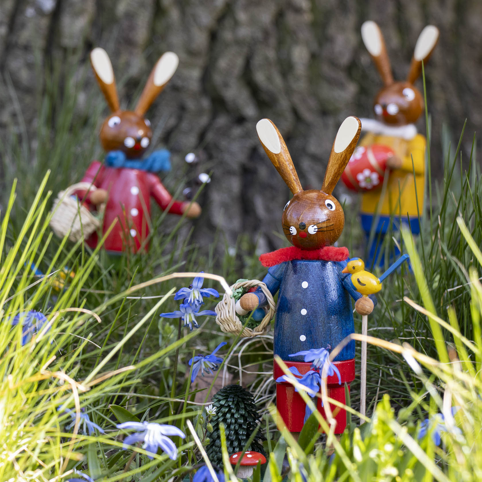 Drei hölzerne Osterhasenfiguren in bunten Farben, die im Gras stehen. Der vordere Hase trägt ein blaues Oberteil, eine rote Hose und hält einen gelben Vogel.