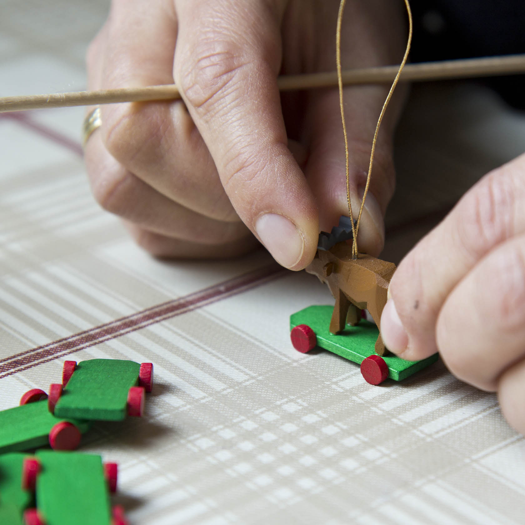 Nahaufnahme von Händen, die ein kleines Holzspielzeug in Form eines Elchs auf roten Rädern bemalen und mit einer Schnur versehen.