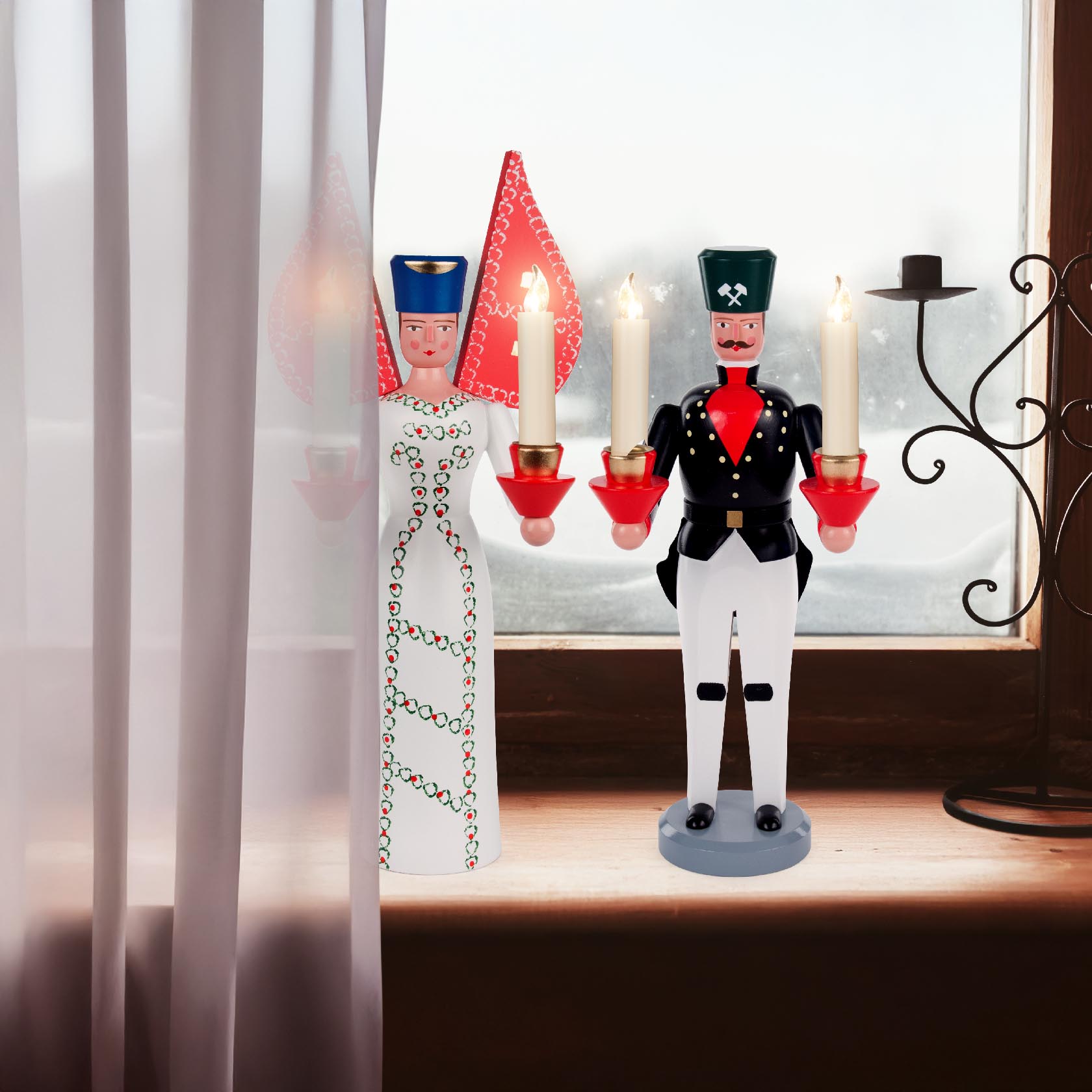 Zwei traditionelle Holzfiguren stehen auf einer Fensterbank. Links ein Engel in weißem Kleid mit roten Verzierungen, rechts ein Bergmann in Tracht, beide halten Kerzen.