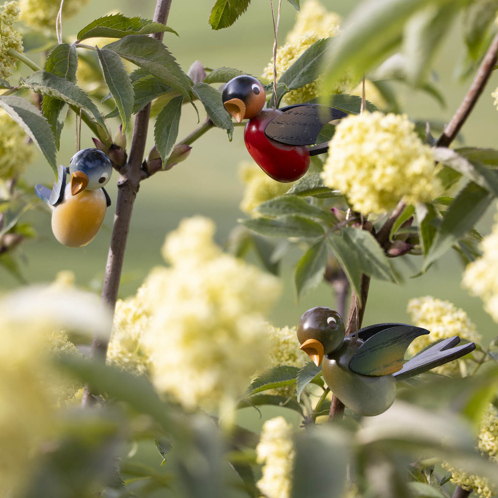 Drei farbenfrohe Vogeldekorationen hängen an einem Strauch mit gelb blühenden Blumen.