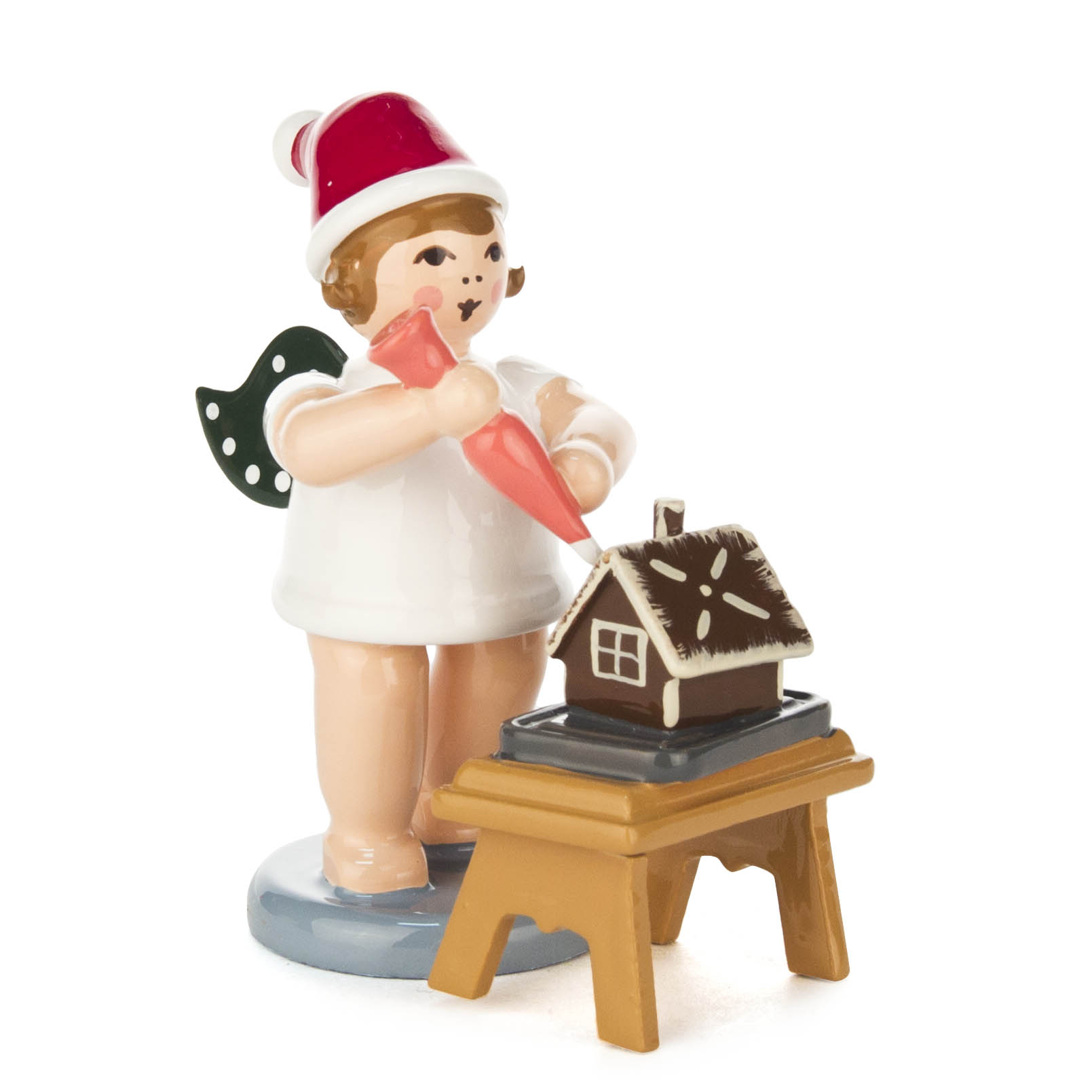 Ein Engel als Bäckerfigur mit grünen Flügeln und roter Mütze hält eine pinke Tortenspritze. Vor ihm steht ein kleines Pfefferkuchenhaus auf einem Tisch. im Dregeno Online Shop günstig kaufen