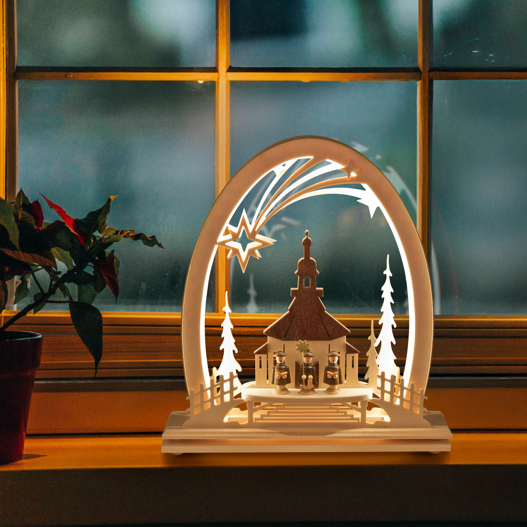 Erzgebirgsbogen mit Seiffener Kirche und Kurrende, beleuchtet durch LED-Lichter. Der Schwibbogen zeigt eine Kirche mit Chorknaben und Sternschnuppe, auf einem Fensterbrett platziert.