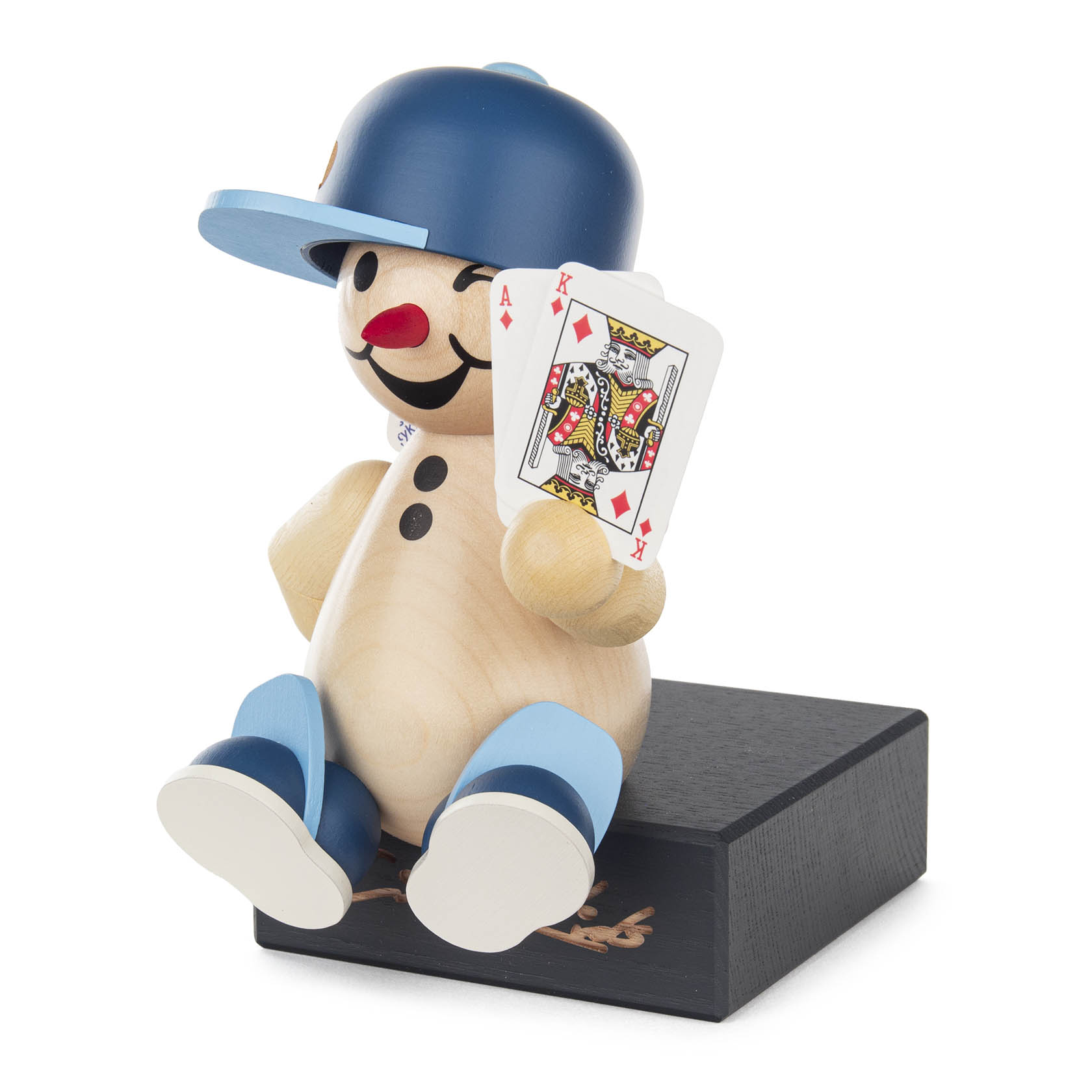Holzfigur eines fröhlichen Jungen mit blauer Kappe, der auf einem schwarzen Sockel sitzt und zwei Spielkarten, Ass und König, in der Hand hält.