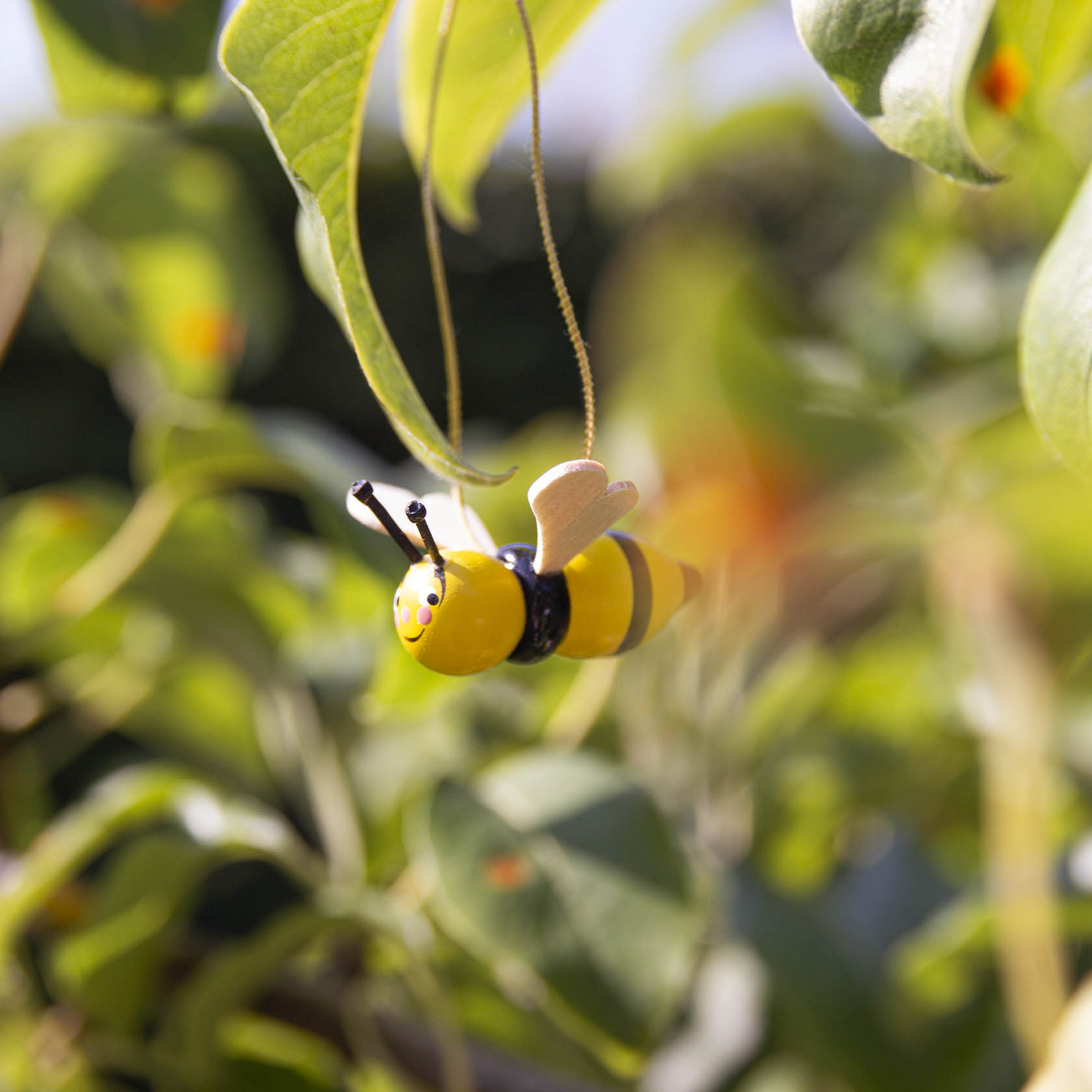 A small, handmade bee made of wood painted yellow with black stripes is hanging from a string among green leaves.