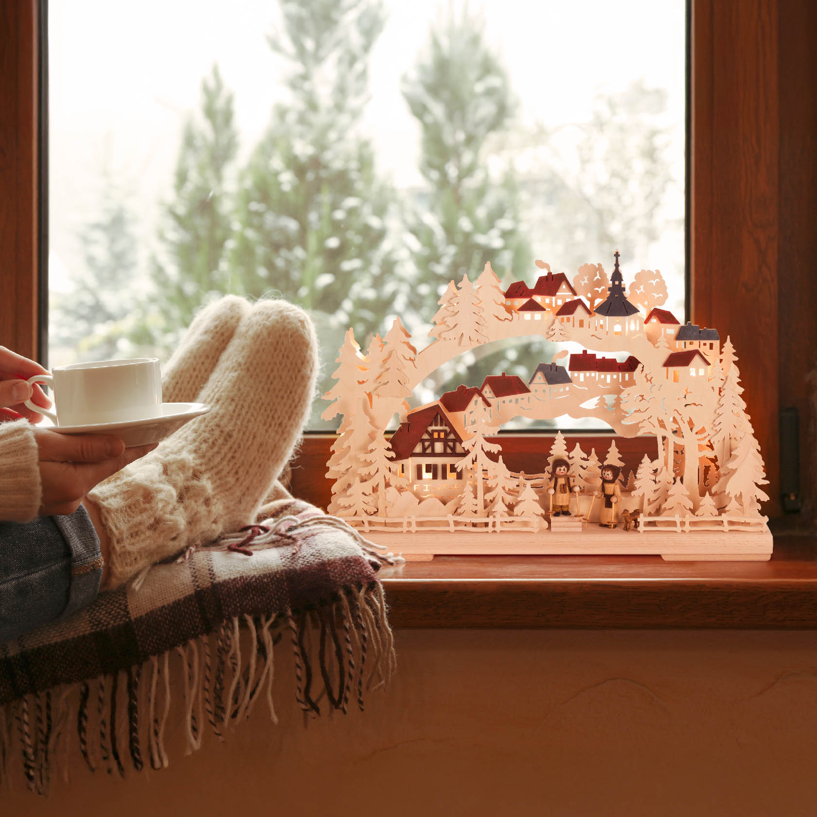 A small illuminated winter scene featuring a Schwibbogen with houses, trees, and a figure, placed on a window sill. A person with wool socks holds a cup.