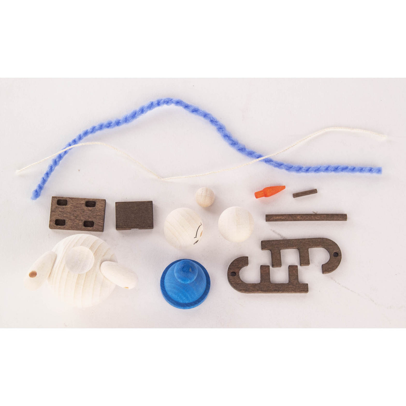 A snowman craft kit including wooden parts for a snowman and sled, a blue base, an orange nose, and blue and white yarn, all laid on a white surface.