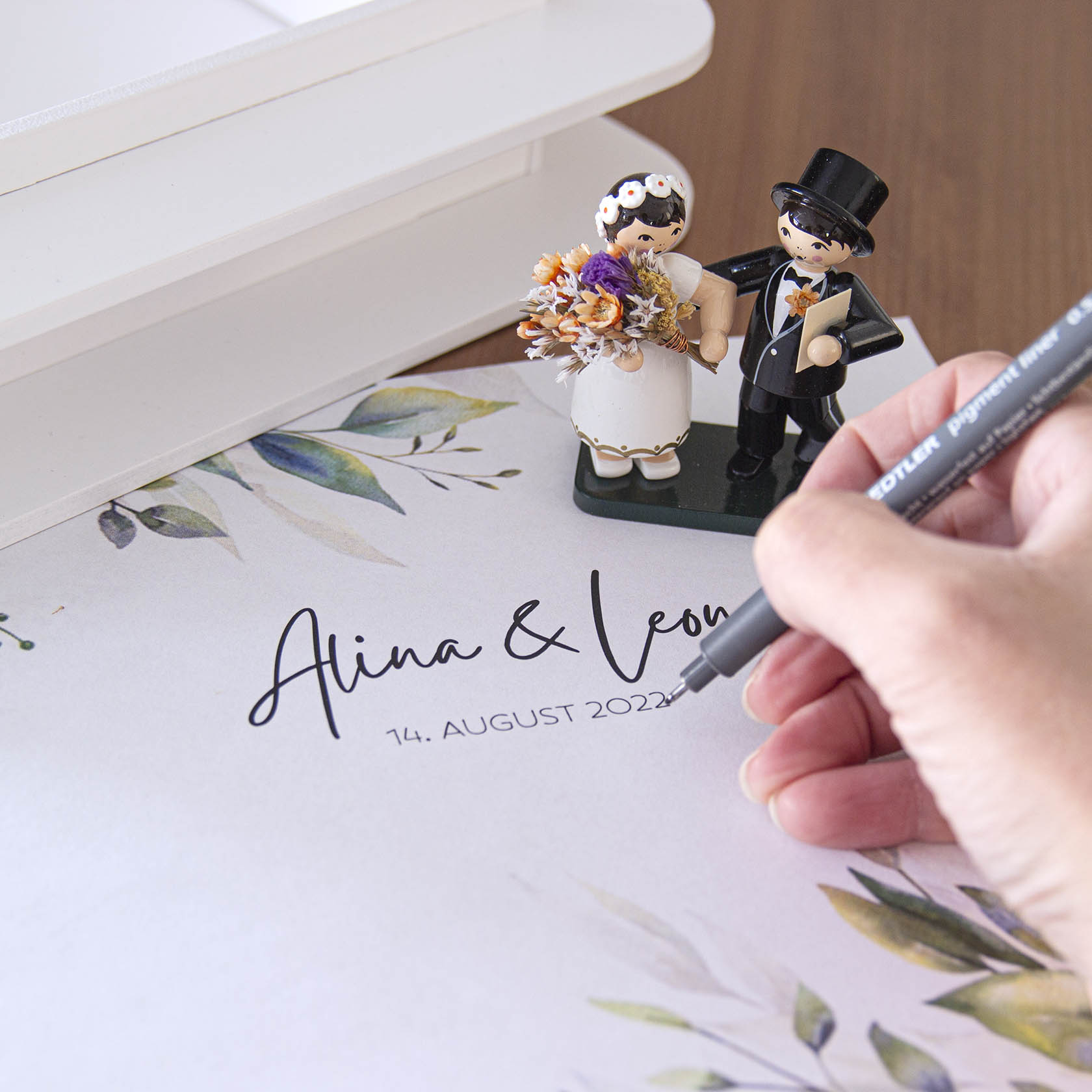 Dekorative Spielfiguren eines Brautpaars auf einem mit Alina & Leon beschrifteten Hochzeitsdokument. Eine Hand schreibt das Datum '14. August 2022'.