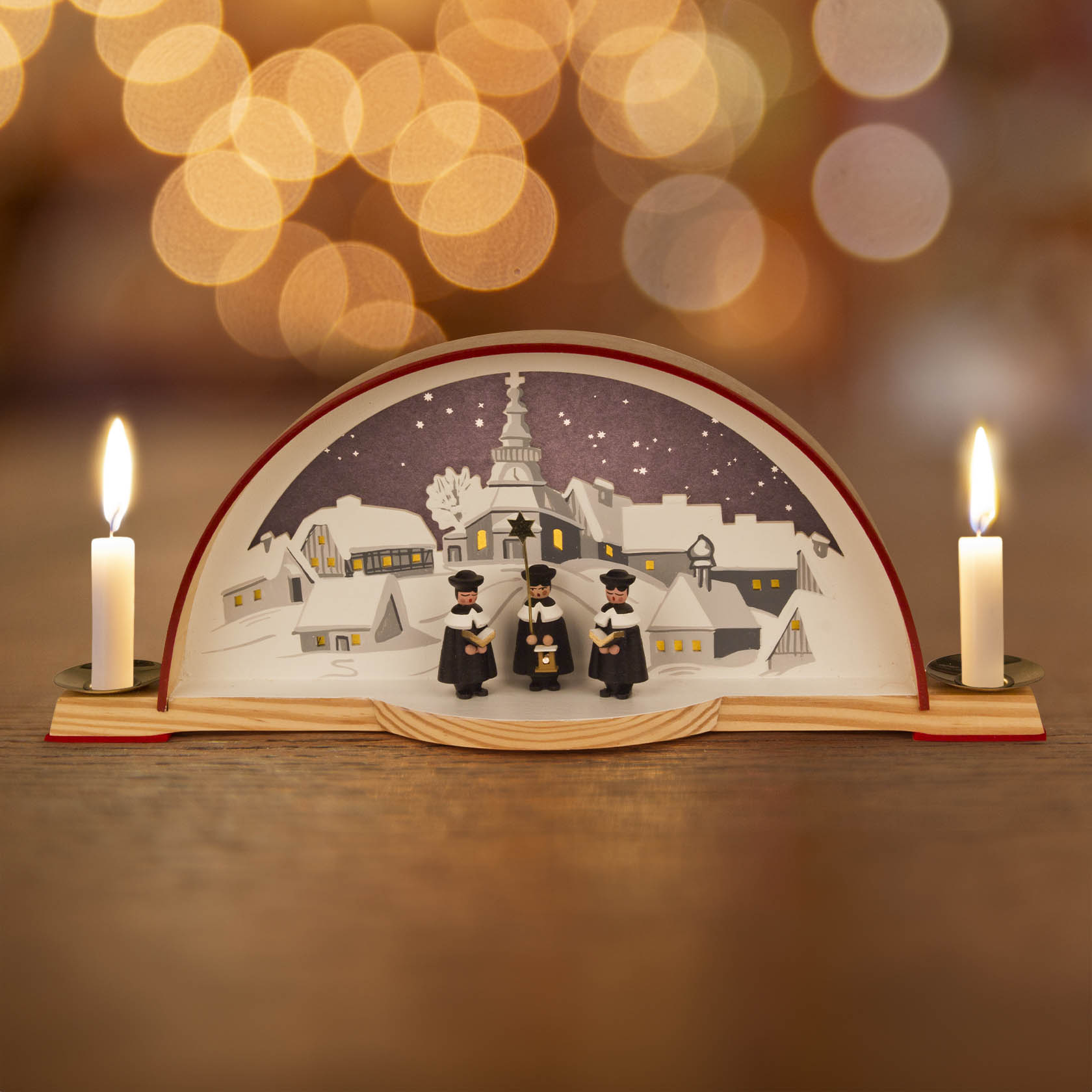 Ein beleuchteter Schwibbogen mit der Seiffener Kirche, drei Kurrendesängern und zwei elektrischen Kerzen. Die Szene zeigt eine Winterlandschaft.