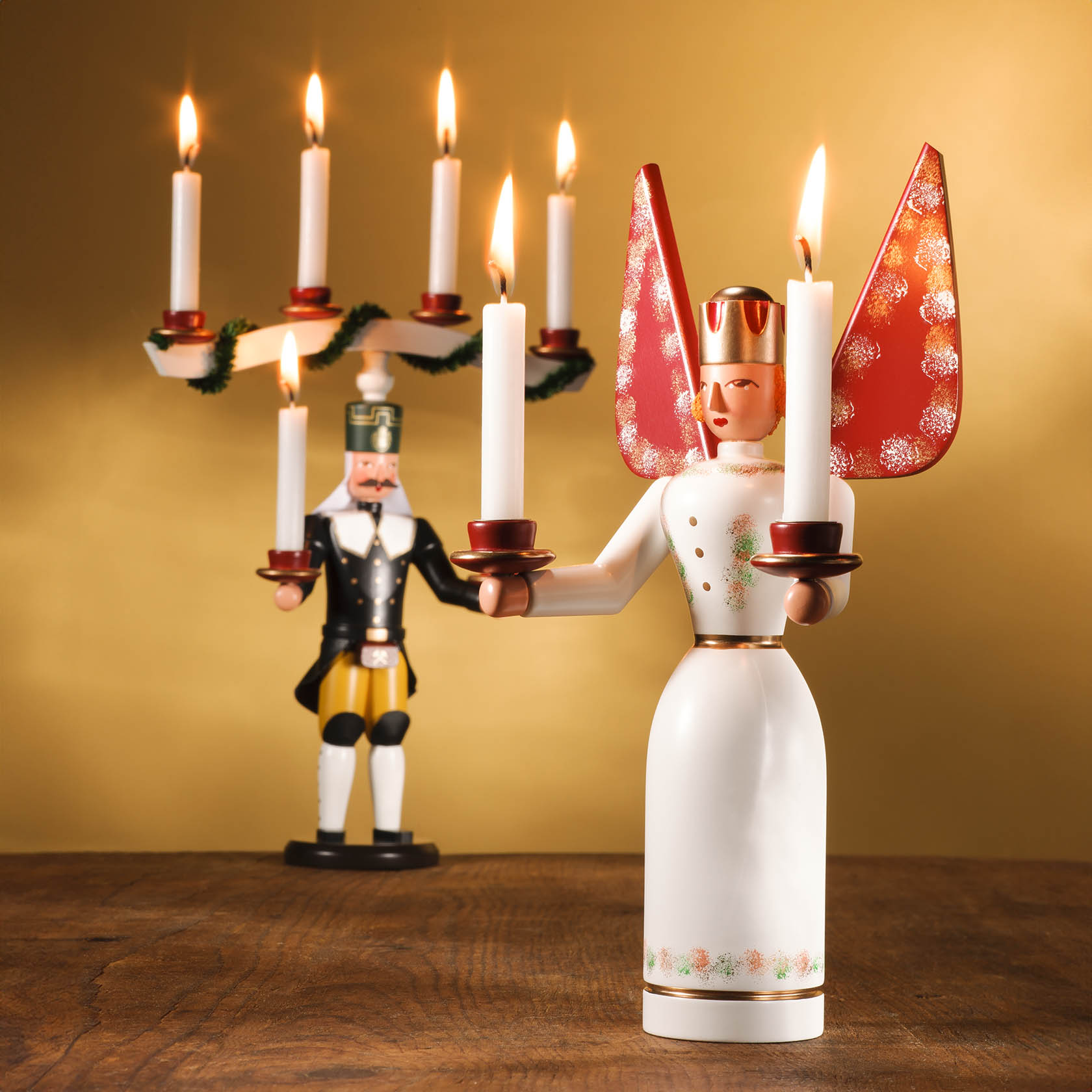 Holzengel mit roten Flügeln und weißen Kerzen in den Händen; im Hintergrund ein Räuchermännchen mit Kerzenhalter.