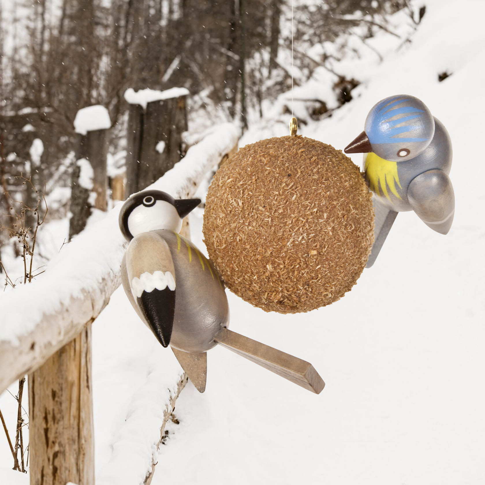 Zwei Holzfiguren von Vögeln, eine Kohlmeise und eine Blaumeise, sitzen auf einem verschneiten Geländer neben einem Meisenknödel.