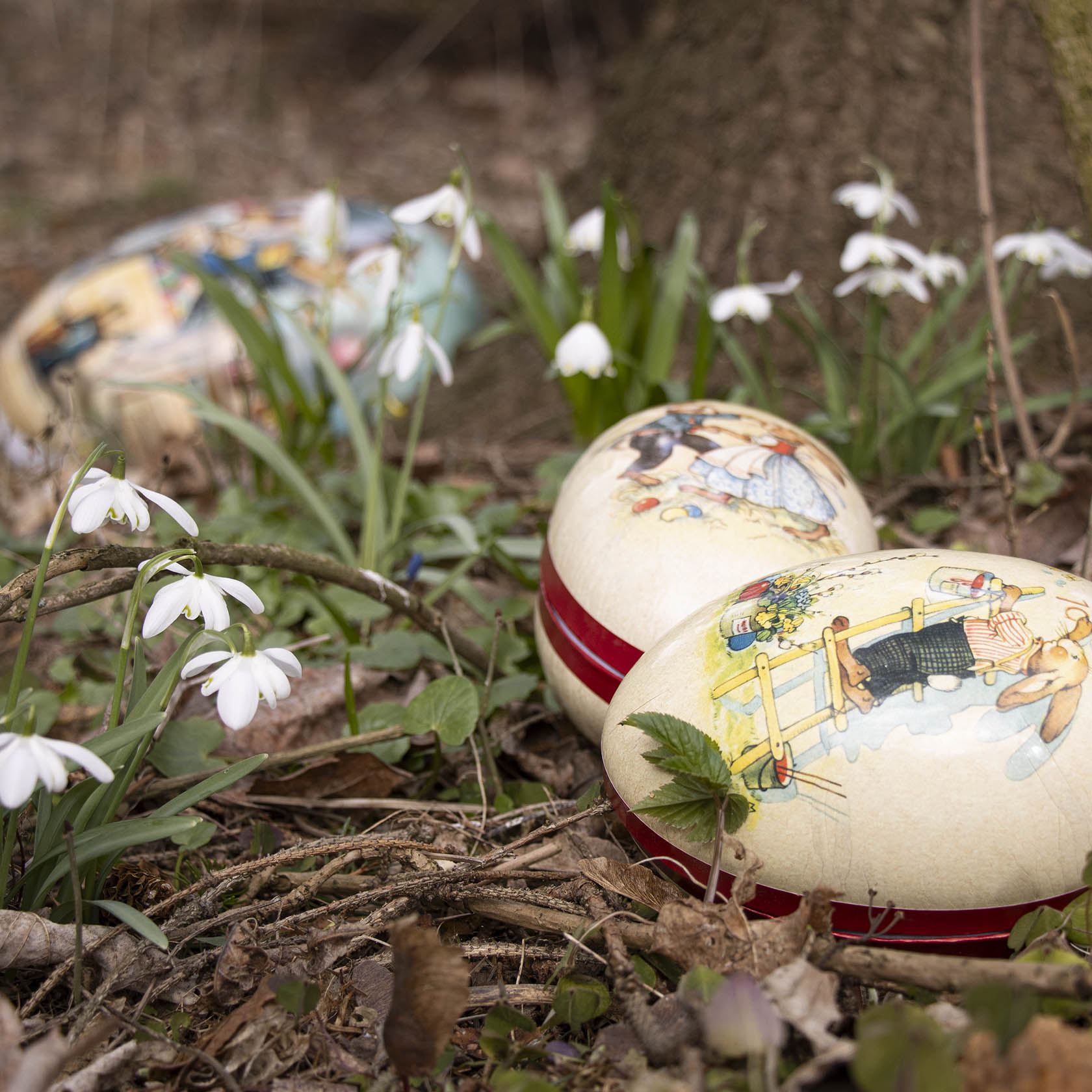 Zwei große Ostereier mit roten Borten und nostalgischen Motiven liegen im Gras neben Schneeglöckchen, im Hintergrund ist ein weiteres Ei zu sehen.