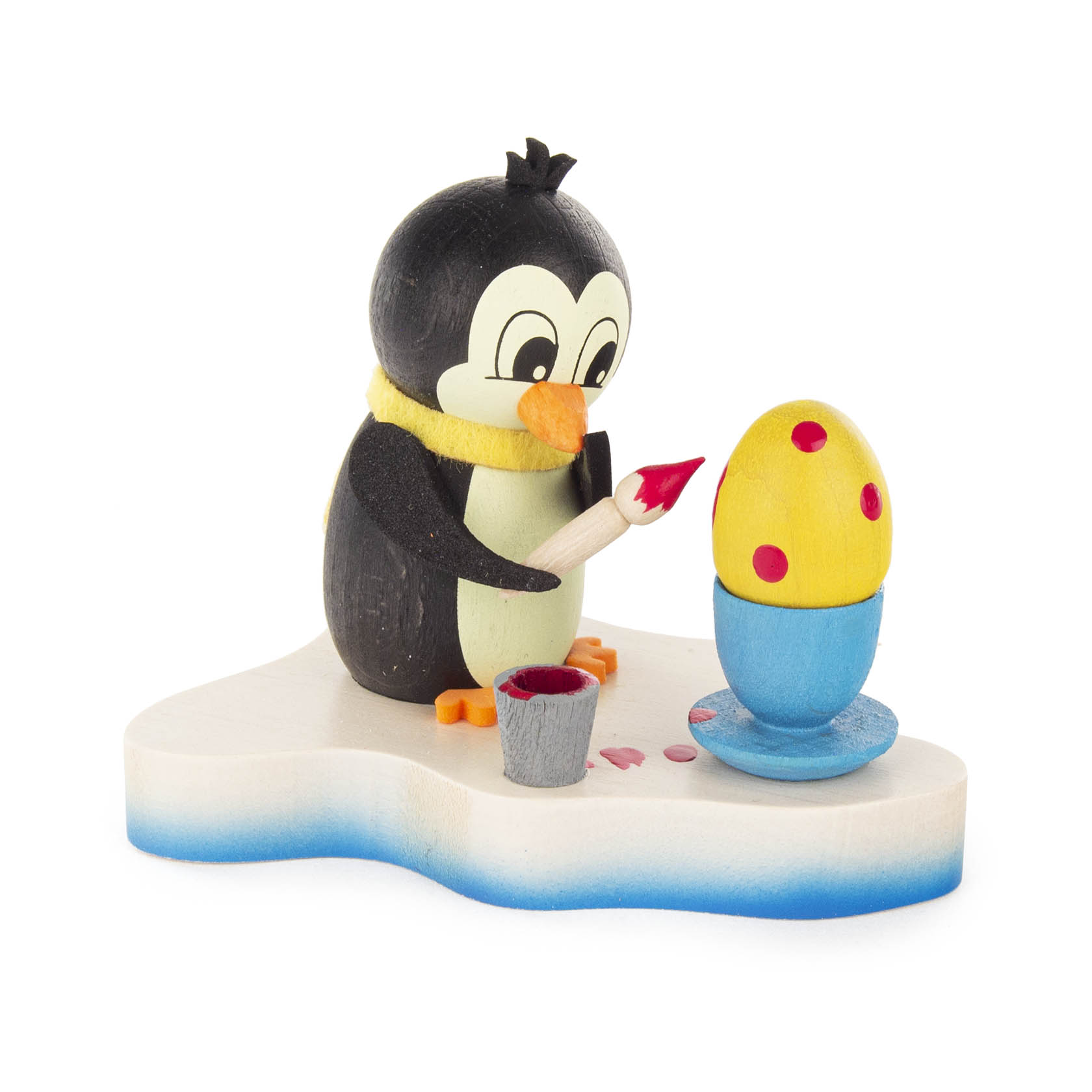 Ein lächelnder Pinguin mit gelbem Schal bemalt mit einem Pinsel ein gelbes Ei mit roten Punkten. Das Ei steht in einem blauen Eierbecher. im Dregeno Online Shop günstig kaufen
