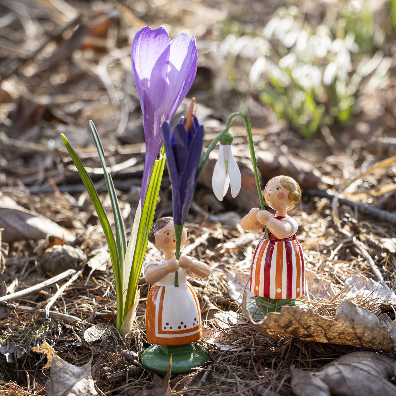 Zwei handbemalte Blumenmädchenfiguren aus Holz, eine hält ein Schneeglöckchen, die andere eine violette Krokusblüte, stehen auf leicht bewachsenem Boden.