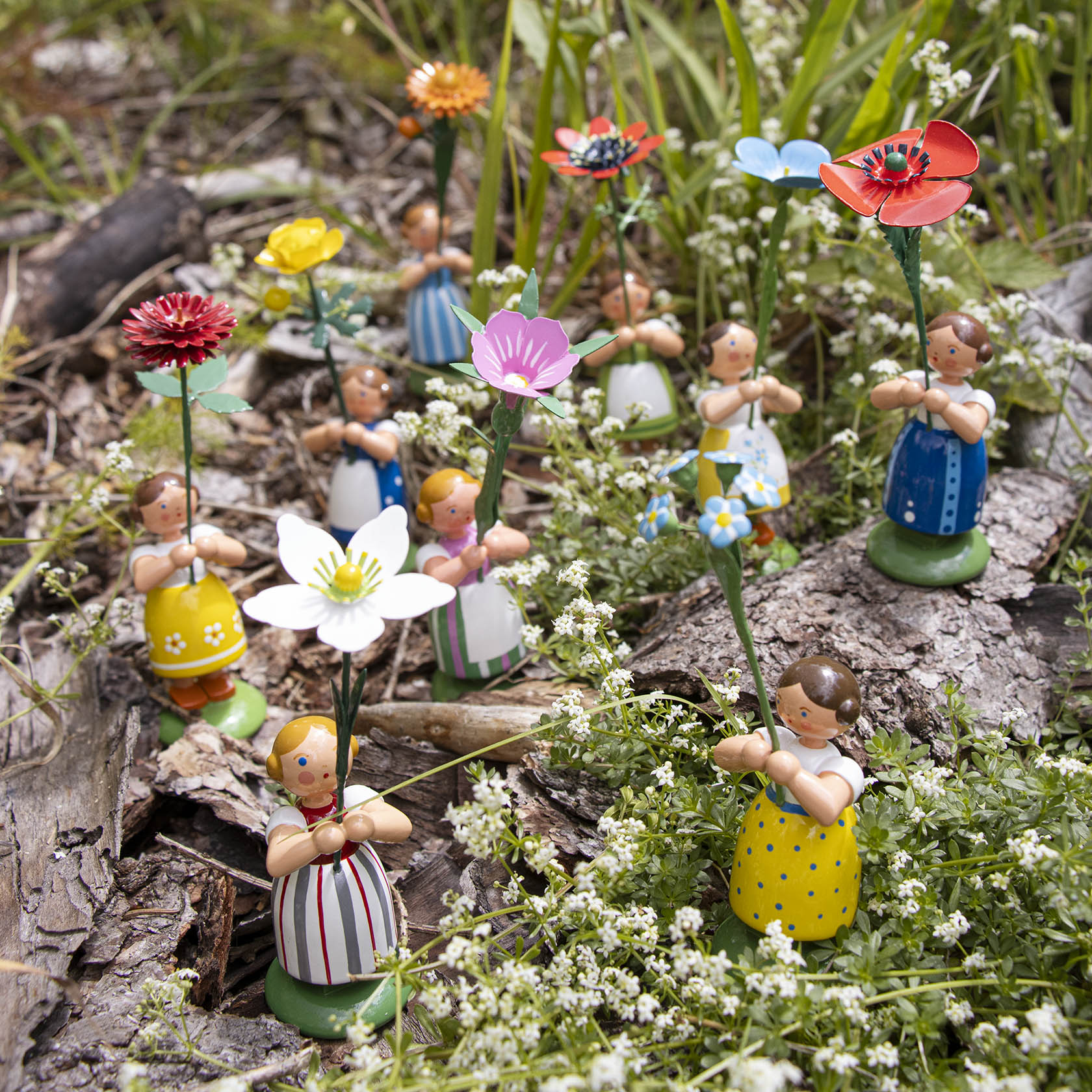 Neun kleine Holzfiguren, als Blumenmädchen gekleidet, jede hält eine Gänseblume aus Holz in der Hand, stehen auf einem bewachsenen Waldboden.